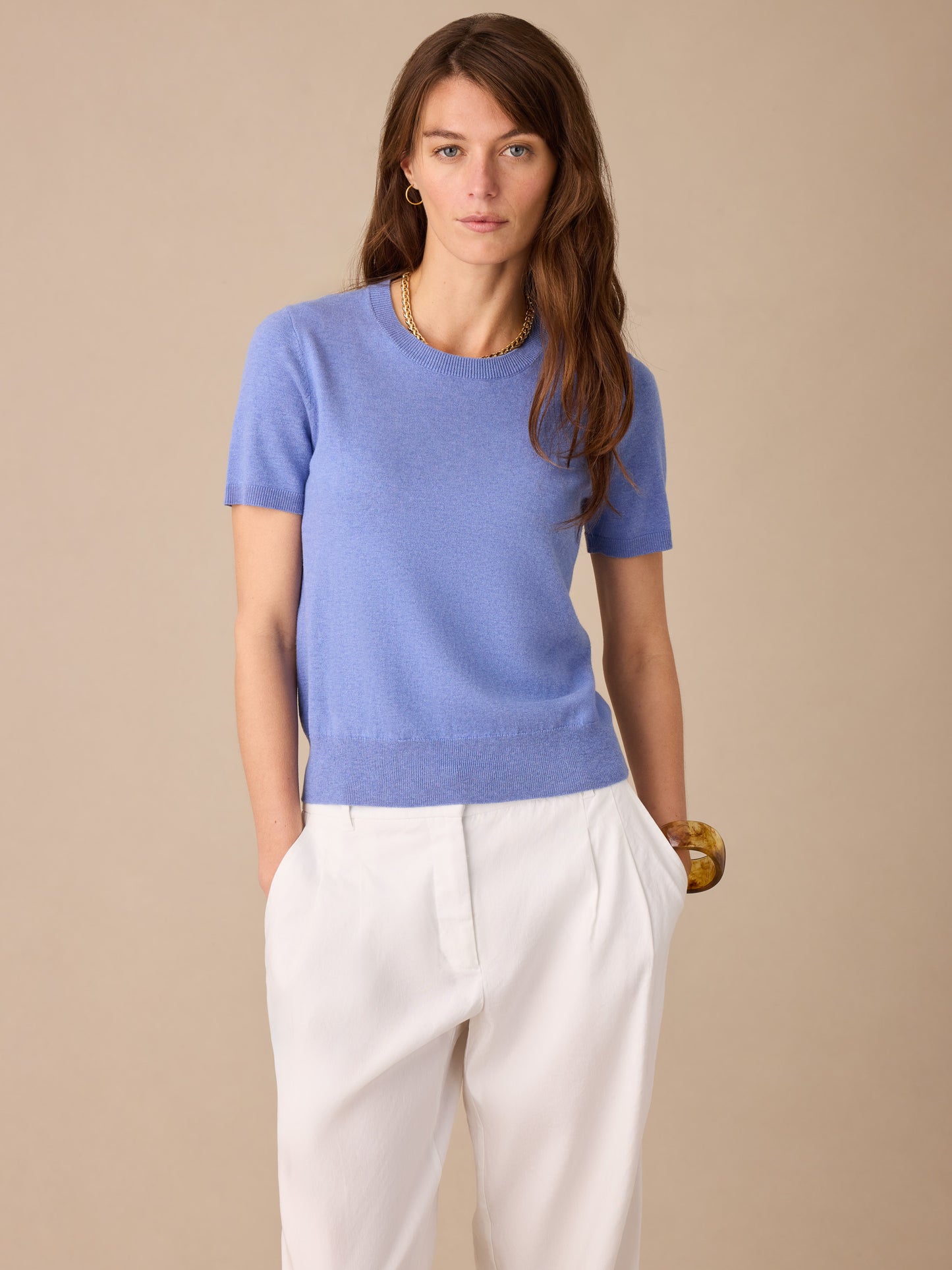 A woman wears the Joelle Sweater by J.McLaughlin in a soft blue hue paired with white pants, showcasing effortlessly stylish, enduring designs and innovative fabrics.