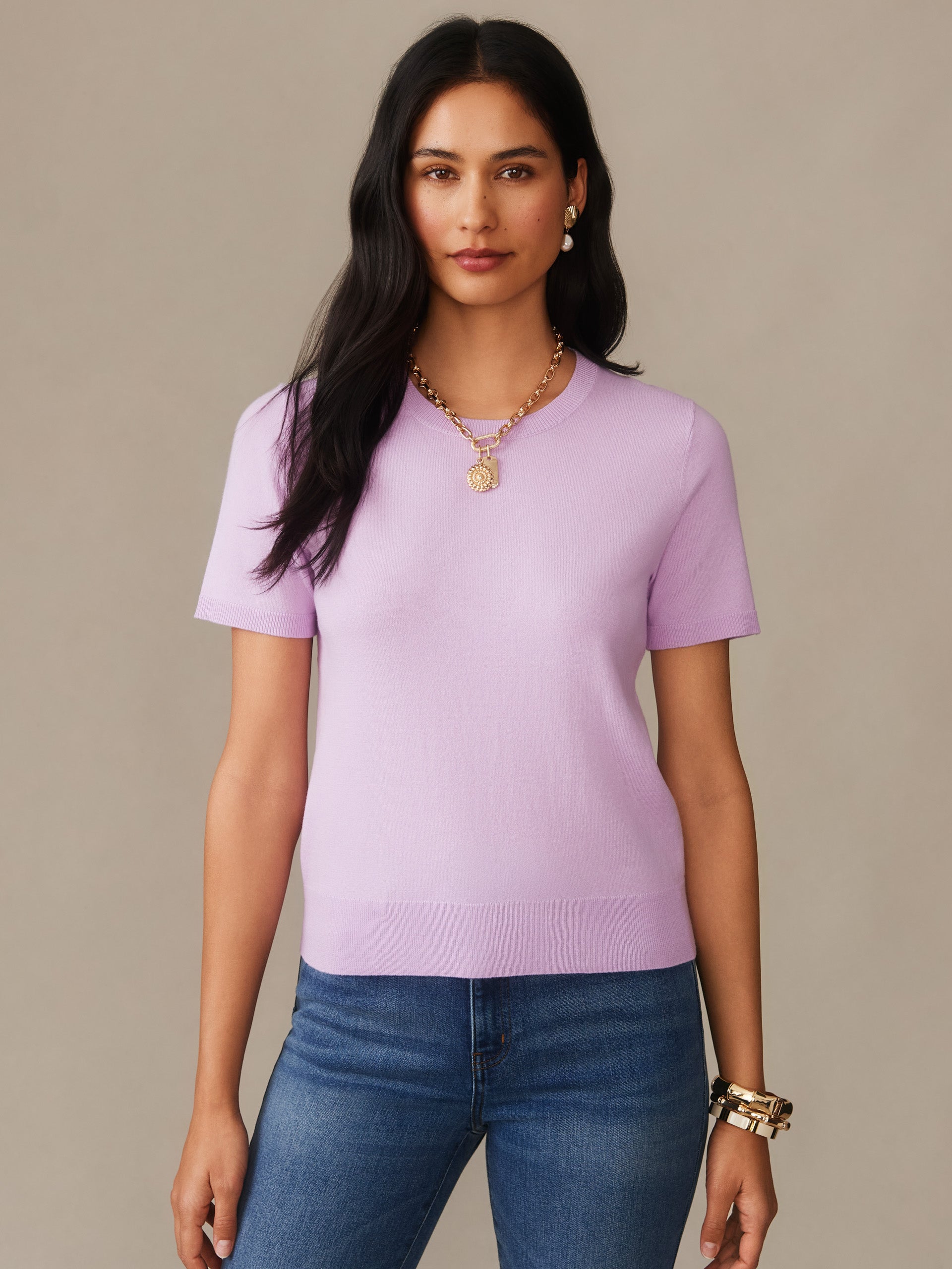 A model wears a lavender Joelle Sweater by J.McLaughlin, paired with blue jeans and gold jewelry, showcasing effortlessly stylish, enduring designs in innovative fabrics.