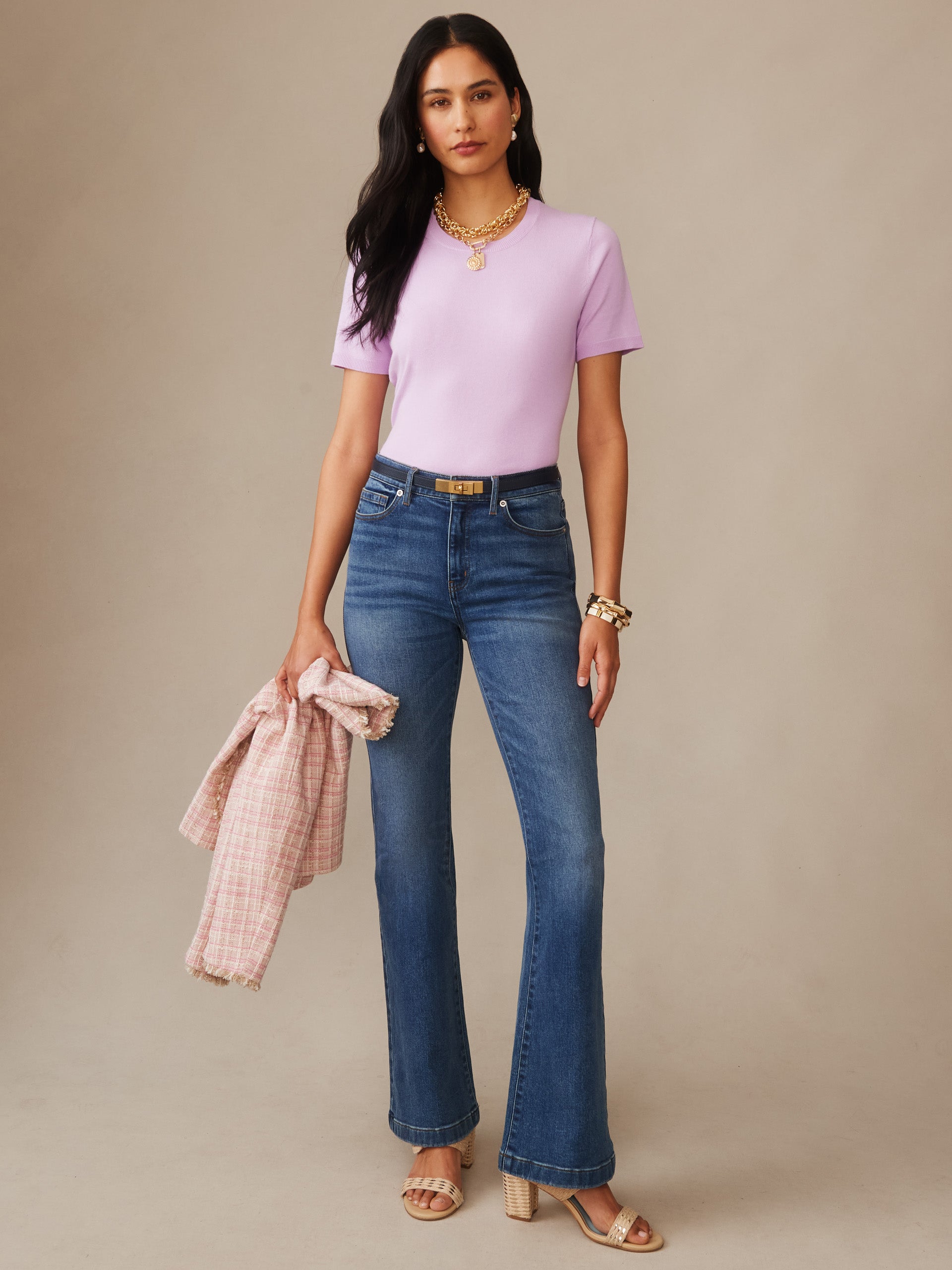Woman wearing a lavender Joelle Sweater by J.McLaughlin paired with blue jeans and gold accessories, holding a pink plaid jacket, showcasing effortlessly stylish, enduring designs for a life well-lived.
