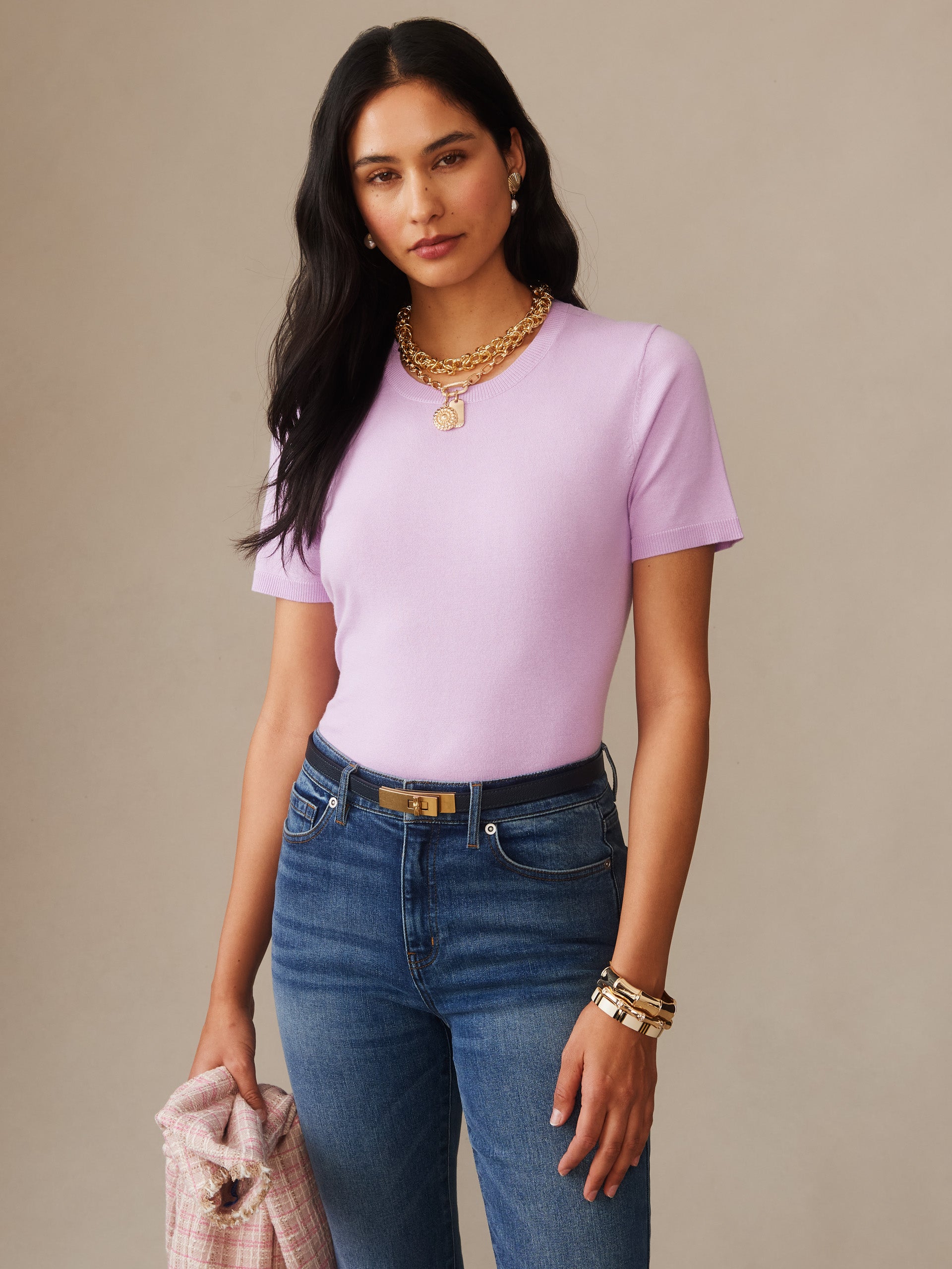 Woman wearing a light purple Joelle Sweater by J.McLaughlin paired with blue jeans and gold jewelry, holding a pink plaid jacket, embodying effortlessly stylish and enduring designs.