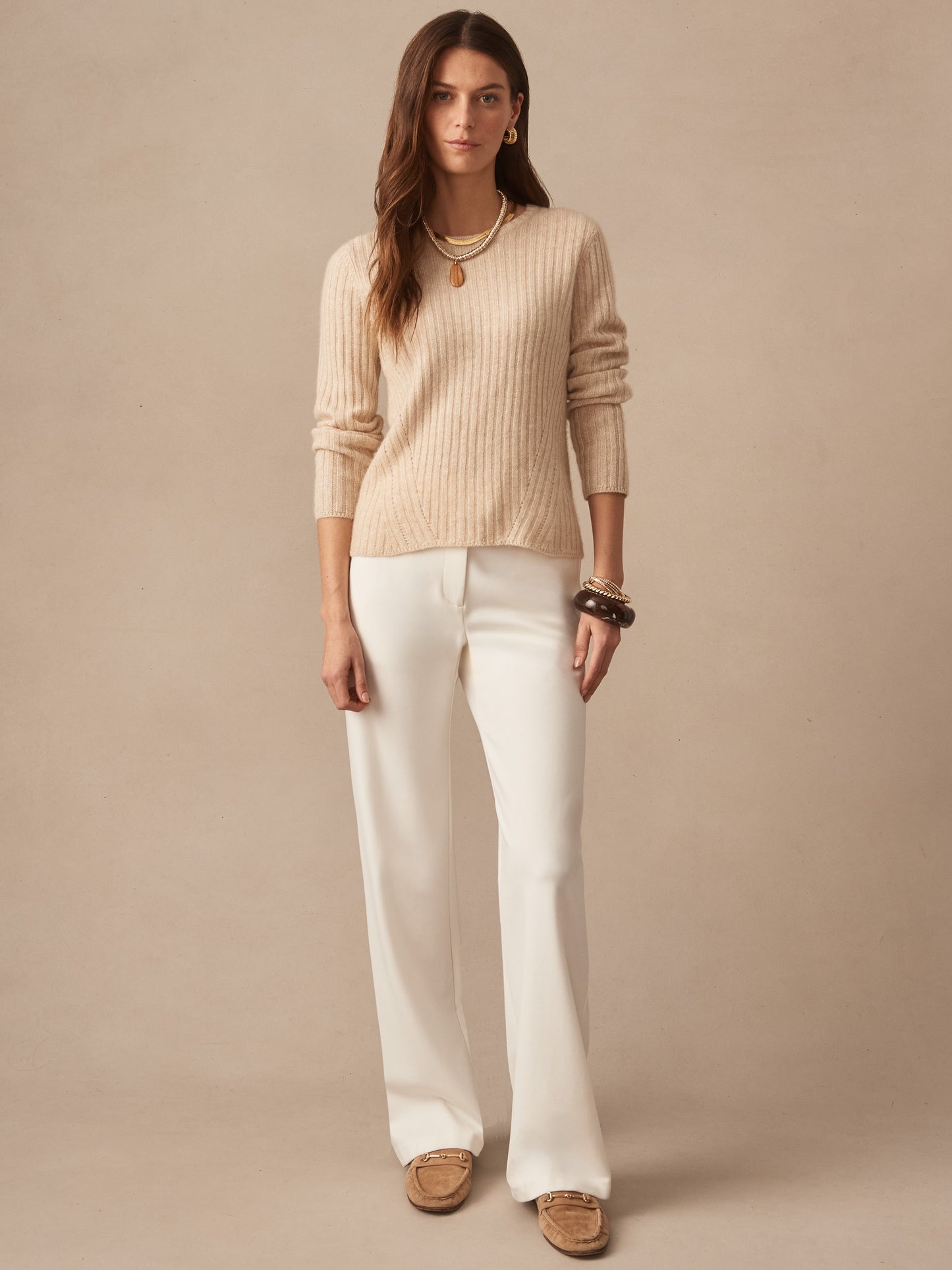Woman wearing a beige Josephine Sweater by J.McLaughlin with white pants and layered necklaces, showcasing effortlessly stylish, enduring designs in a timeless setting.