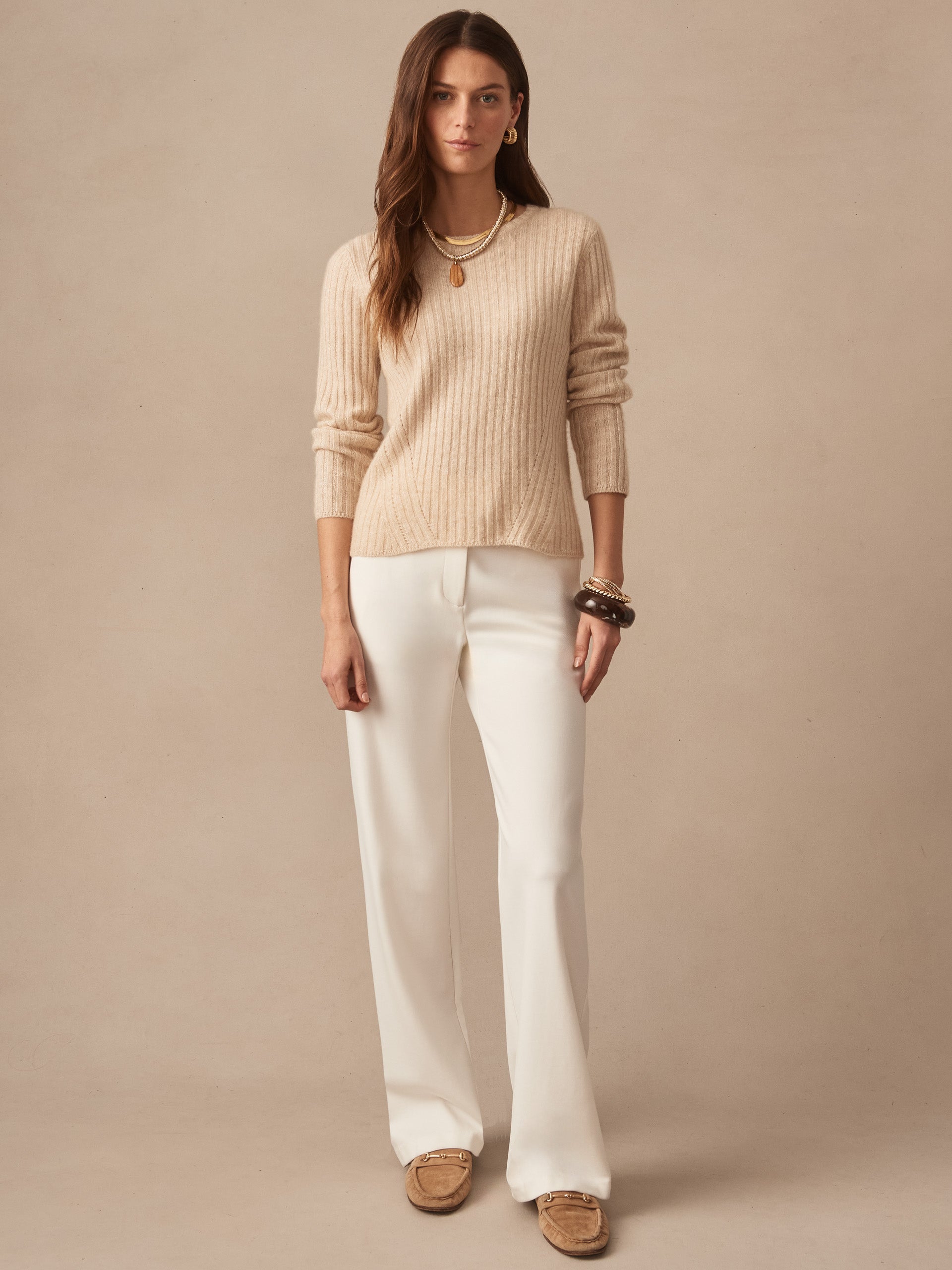 Woman wearing a beige Josephine Sweater by J.McLaughlin with white pants and layered necklaces, showcasing effortlessly stylish, enduring designs in a timeless setting.