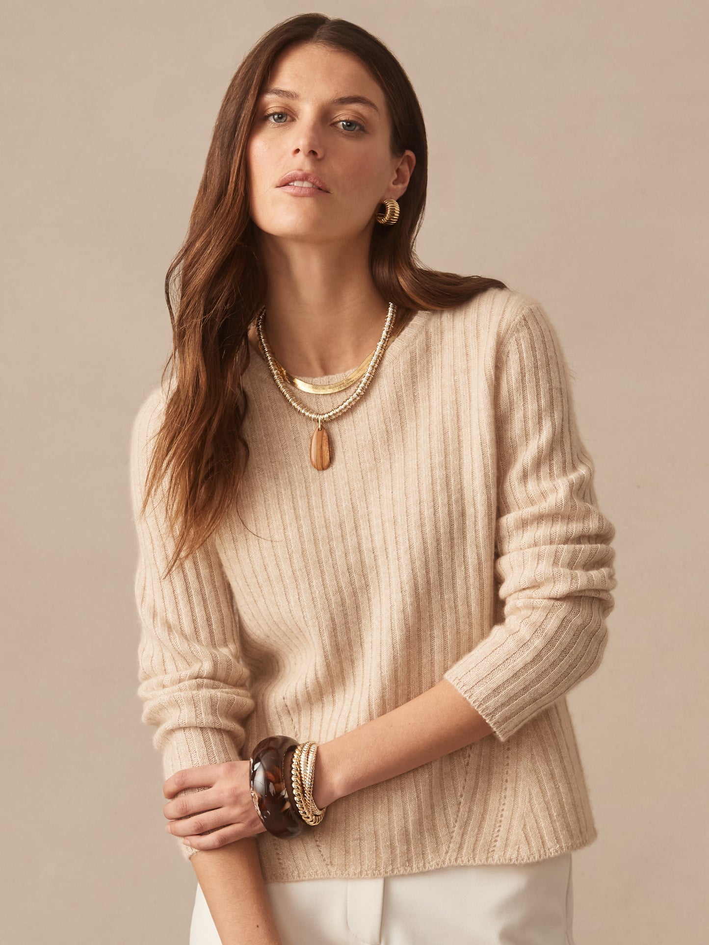 Woman wearing a soft oatmeal-colored Josephine Sweater by J.McLaughlin, styled with layered necklaces and bracelets, showcasing effortlessly stylish, enduring designs in a timeless look.