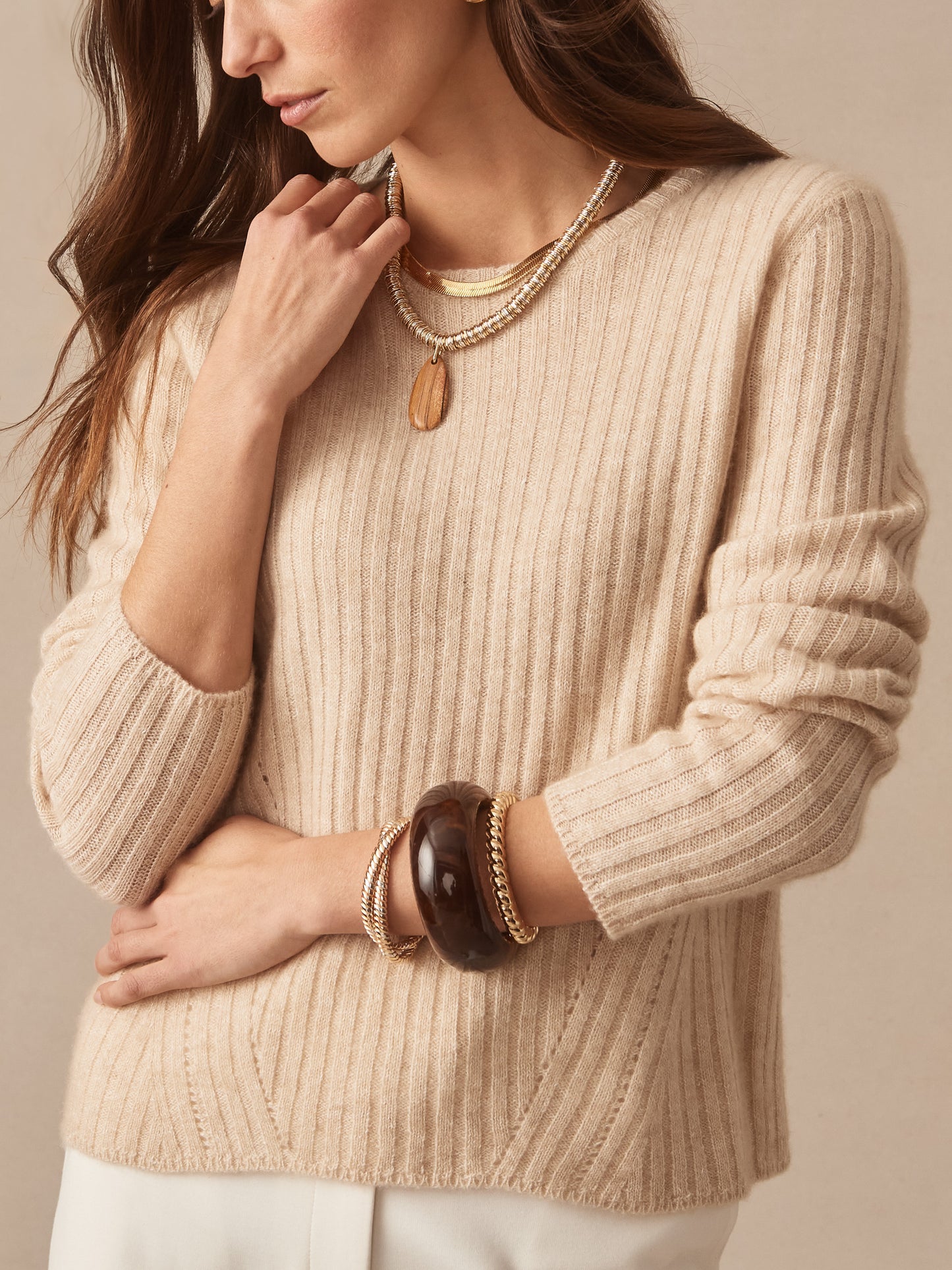 A close-up of a woman wearing a ribbed oatmeal-colored Josephine Sweater by J.McLaughlin, styled with layered necklaces and bracelets, showcasing effortlessly stylish, enduring designs and innovative fabrics for a destination of defining style. Life, Well-Lived.