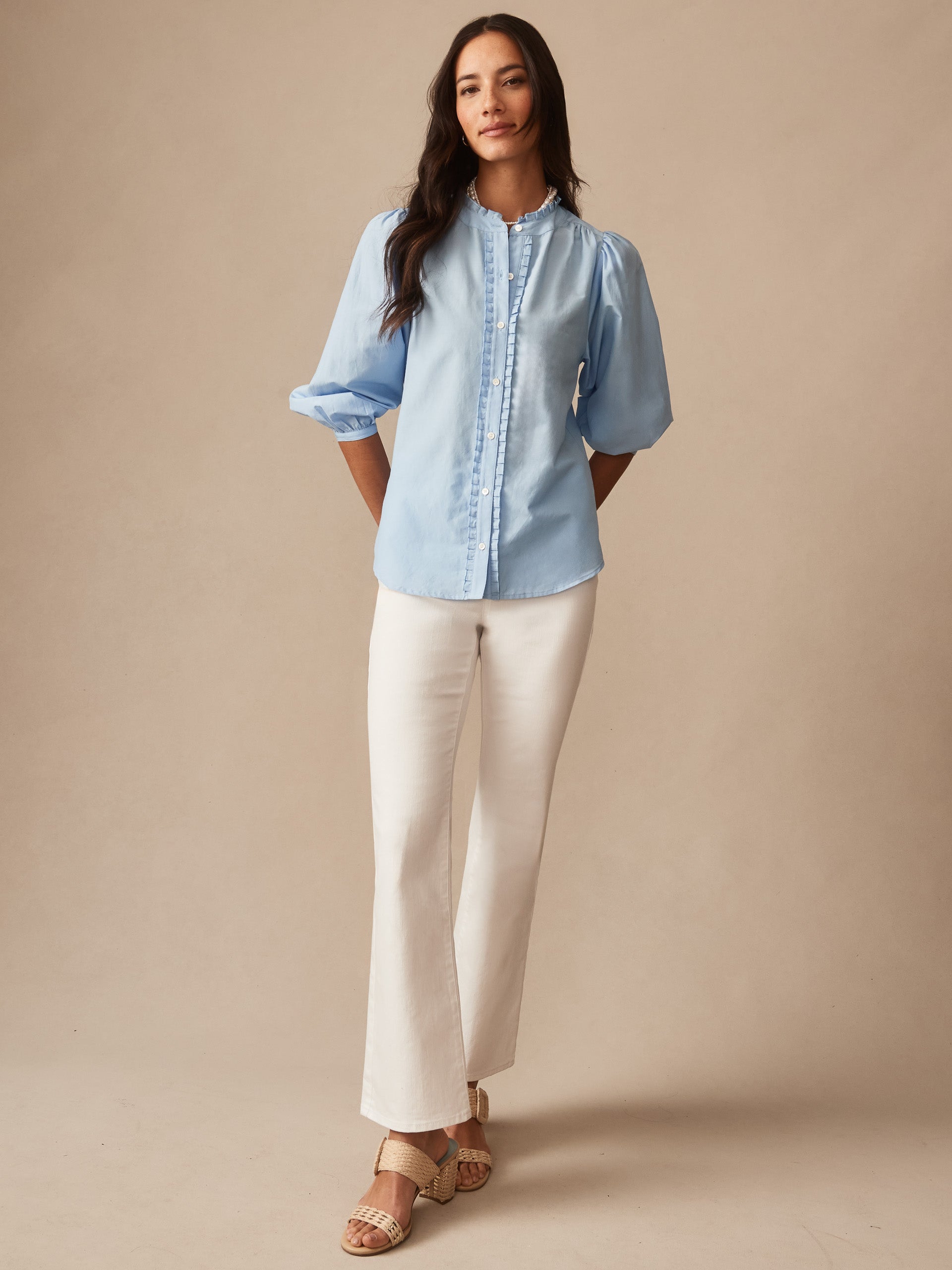 Woman wearing a light blue Josette Blouse by J.McLaughlin paired with white pants and sandals, showcasing effortlessly stylish and enduring designs with a legacy of timeless fashion.