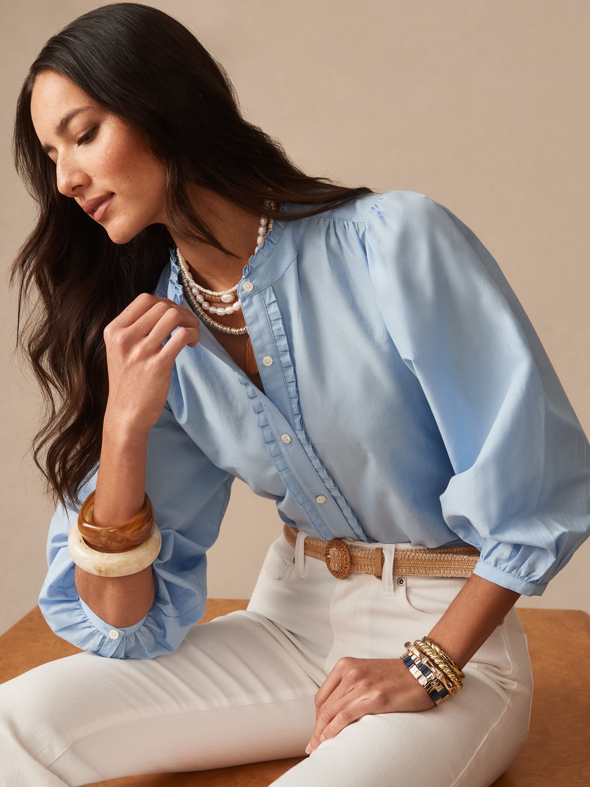 Woman wearing a light blue Josette Blouse by J.McLaughlin, styled with layered pearl necklaces, white pants, and mixed bracelets, embodying effortlessly stylish and enduring designs.