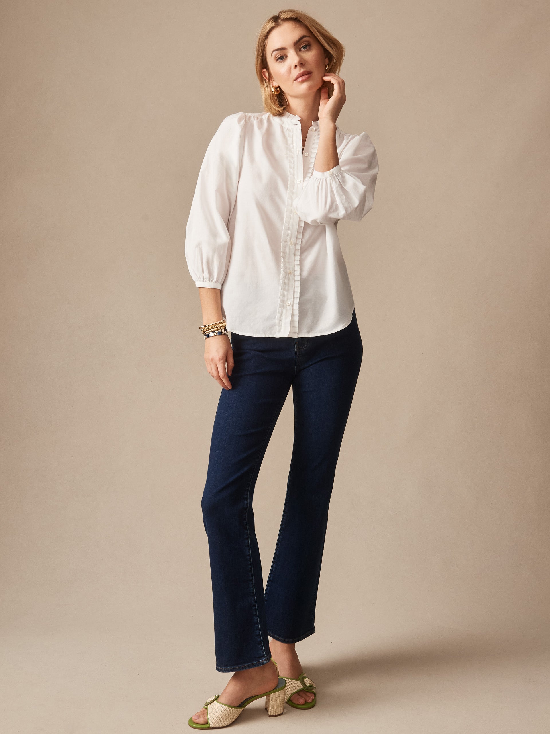 A stylish woman wearing dark blue Elvira Jeans by J.McLaughlin paired with a crisp white blouse and green block-heeled sandals, embodying effortlessly stylish, enduring designs.