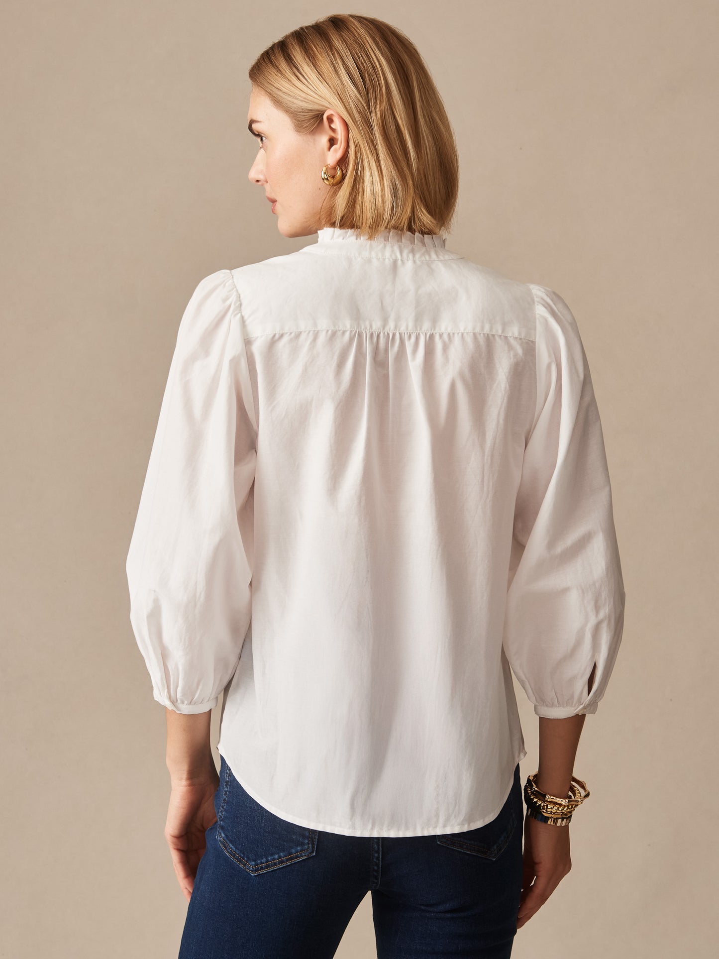 Woman wearing a white Josette Blouse by J.McLaughlin, featuring puffed sleeves and ruffled collar, paired with dark jeans, showcasing effortlessly stylish, enduring designs from a legacy of timeless fashion. A destination for defining style. Life, Well-Lived.