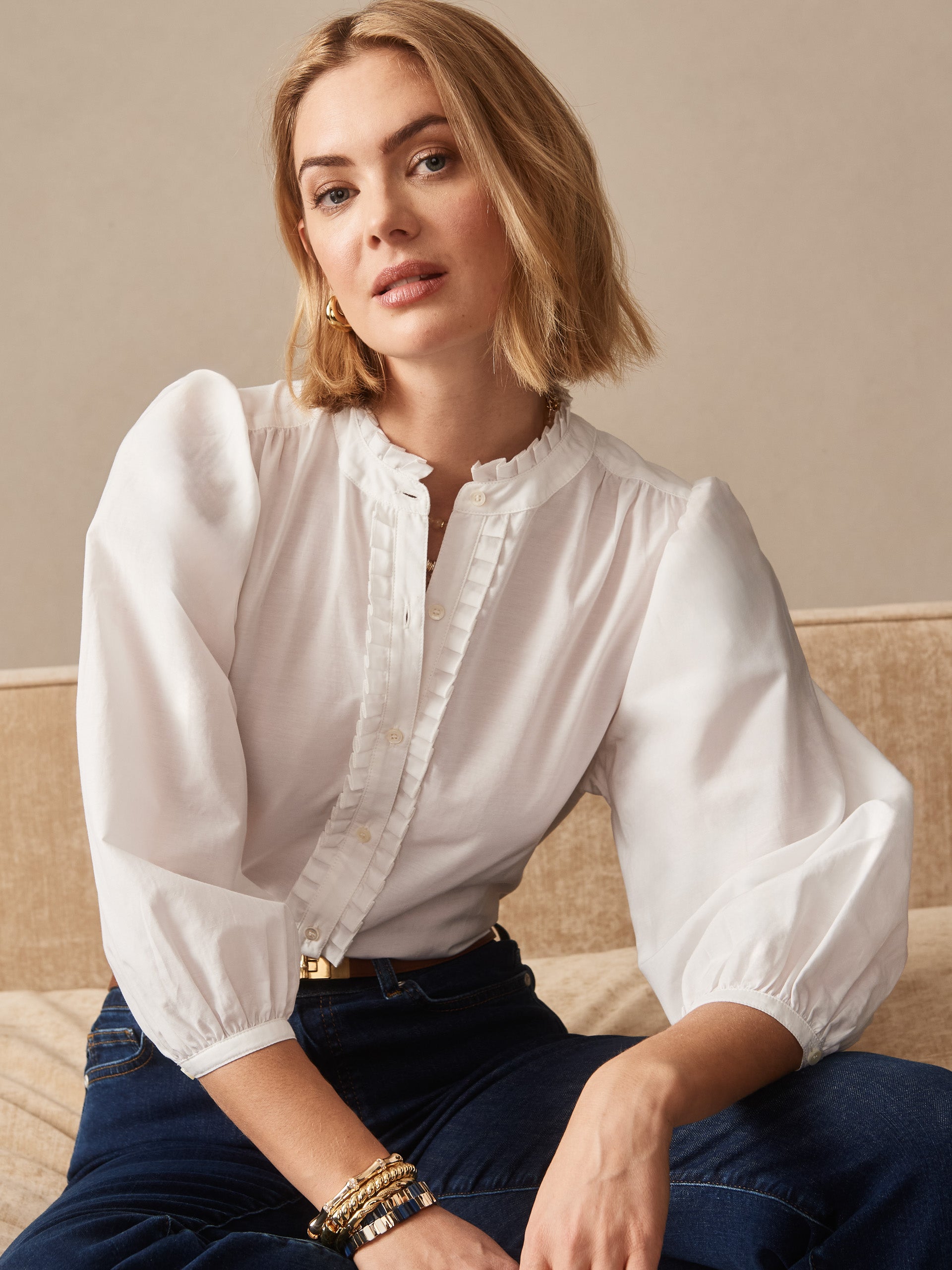 Female model wearing a white Josette Blouse with puffed sleeves and ruffled collar, paired with dark jeans, showcasing J.McLaughlin’s effortlessly stylish, enduring designs and legacy of timeless fashion.