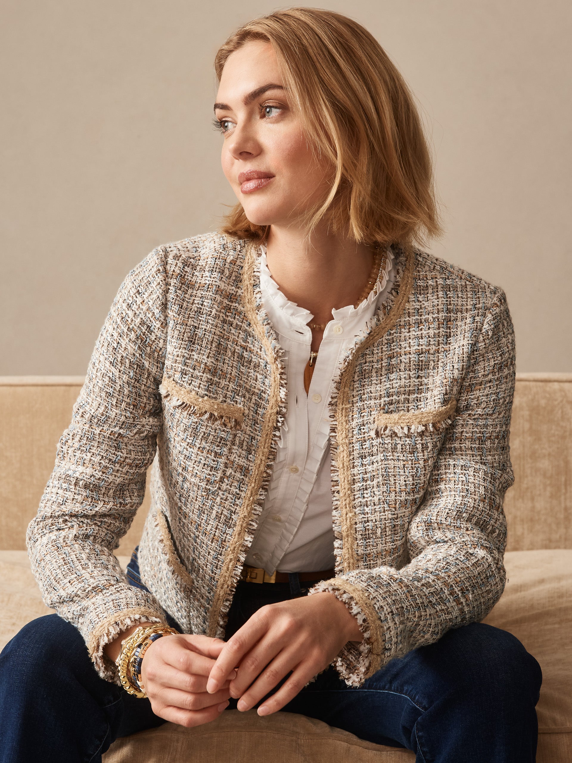 Woman seated on a beige couch wearing the Martha Tweed Jacket by J.McLaughlin over a white blouse and dark jeans, showcasing effortlessly stylish, enduring designs in a classic tweed fabric.