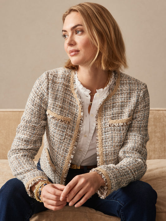 Woman seated on a beige couch wearing the Martha Tweed Jacket by J.McLaughlin over a white blouse and dark jeans, showcasing effortlessly stylish, enduring designs in a classic tweed fabric.
