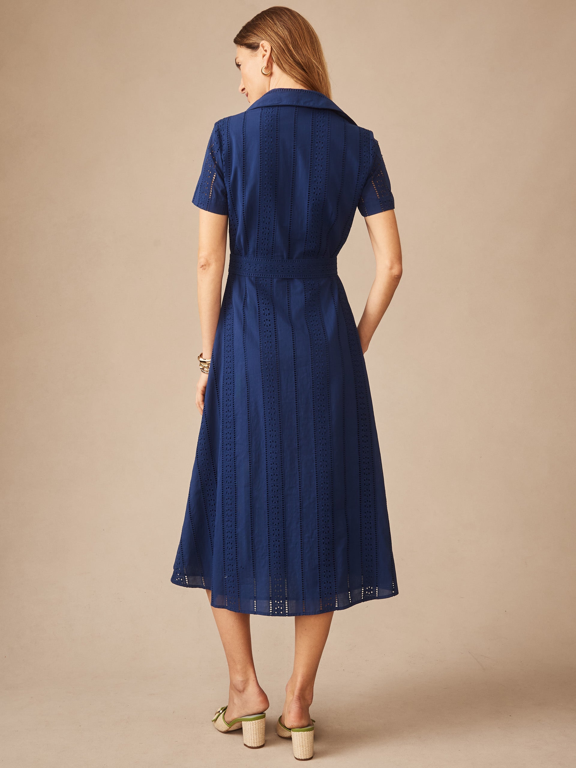 A woman models the Kalinda Dress in Eyelet by J.McLaughlin, featuring a navy blue, eyelet-patterned fabric with a classic collar and short sleeves, embodying effortlessly stylish, enduring designs and innovative fabrics in a legacy of timeless fashion. A destination for defining style. Life, Well-Lived.