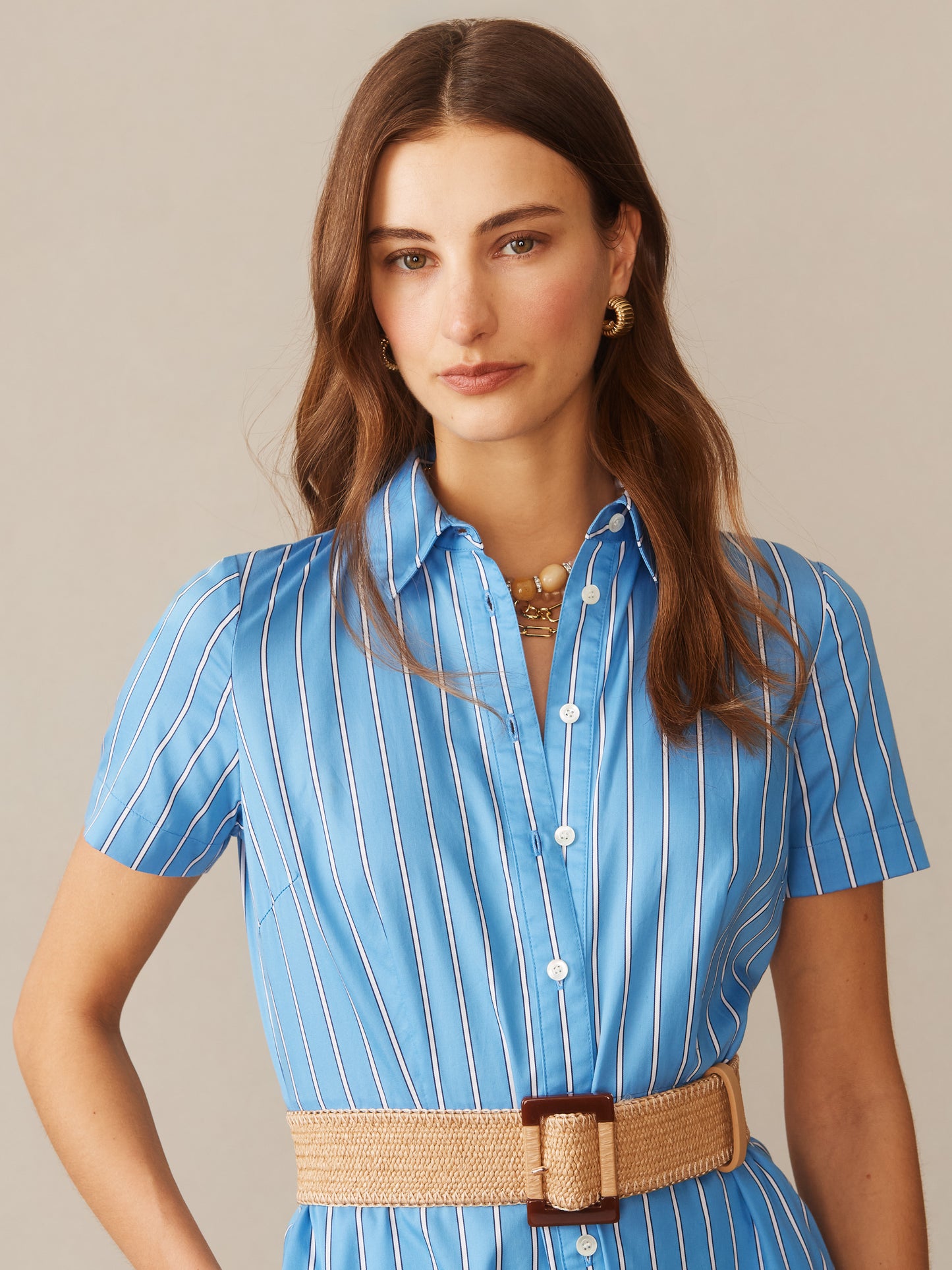 Woman wearing a blue and white vertically striped Kalinda Dress in Stripe by J.McLaughlin, styled with a woven belt and gold hoop earrings, showcasing effortlessly stylish and enduring designs.