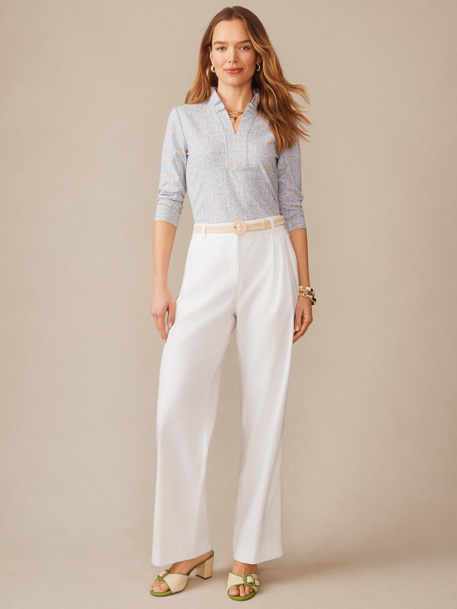 A woman wearing the Kate Ruffle Top in Bamboo Lane by J.McLaughlin paired with white wide-leg pants and green open-toe shoes, showcasing effortlessly stylish, enduring designs crafted from innovative fabrics. A destination for defining style. Life, Well-Lived.