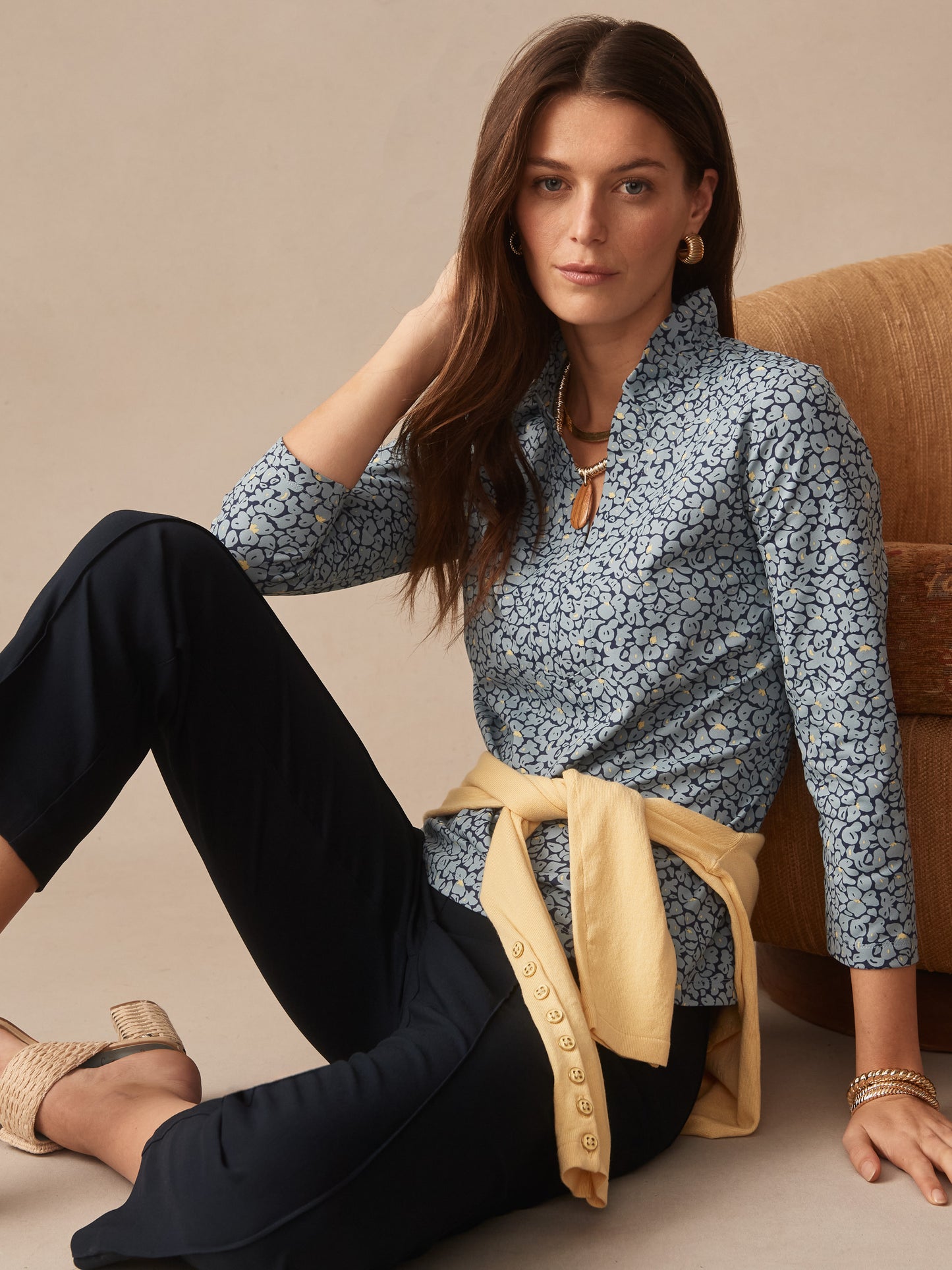 Woman seated wearing the Kate Ruffle Top in Celandine by J.McLaughlin, styled with navy pants and a light yellow sweater tied at the waist, showcasing effortlessly stylish, enduring designs and innovative fabrics.
