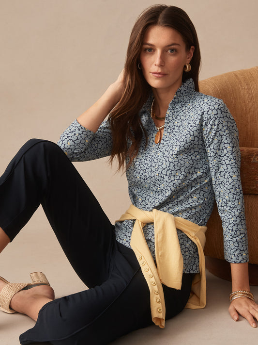 Woman seated wearing the Kate Ruffle Top in Celandine by J.McLaughlin, styled with navy pants and a light yellow sweater tied at the waist, showcasing effortlessly stylish, enduring designs and innovative fabrics.