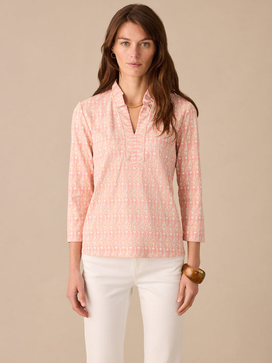 Woman wearing a pink patterned Kate Ruffle Top in Neo Jewel Ikat by J.McLaughlin, styled with white pants and a wooden bracelet, showcasing effortlessly stylish design and innovative fabrics.
