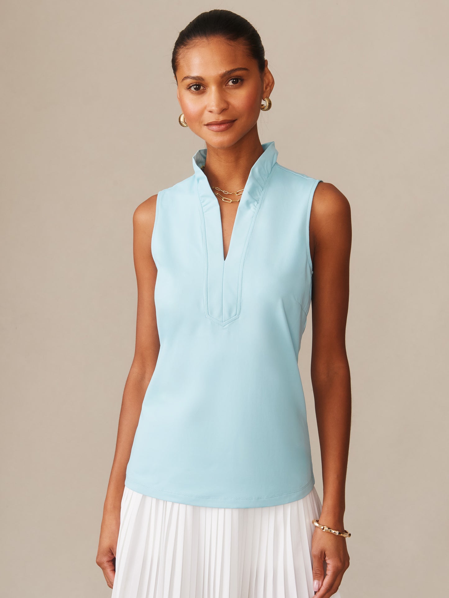 Woman wearing a tropical blue Kate Sleeveless Ruffle Top by J.McLaughlin, showcasing an effortlessly stylish, enduring design paired with a white pleated skirt, embodying a legacy of timeless fashion and innovative fabrics.