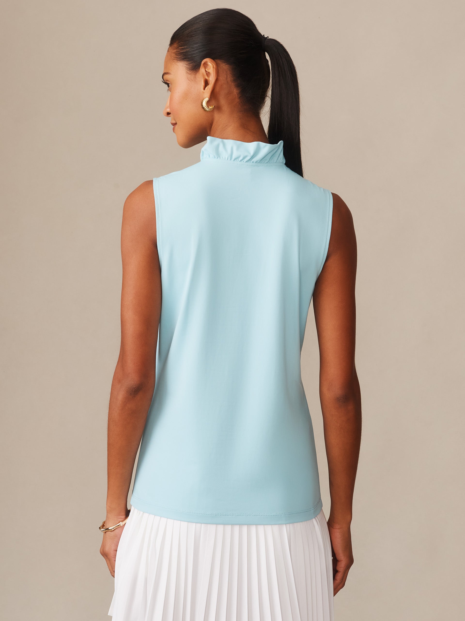 Woman wearing a tropical blue Kate Sleeveless Ruffle Top by J.McLaughlin, styled with a white pleated skirt, showcasing effortlessly stylish, enduring designs and innovative fabrics.