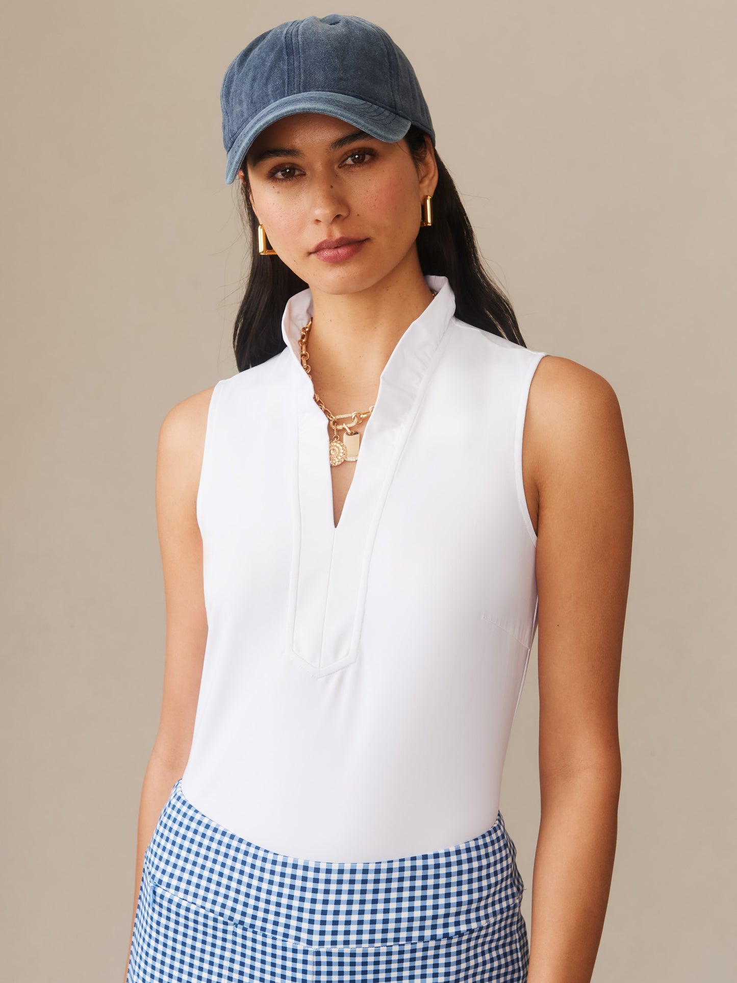 Woman wearing a white Kate Sleeveless Ruffle Top by J.McLaughlin paired with blue gingham skirt and a casual blue cap, showcasing effortlessly stylish, enduring designs and innovative fabrics.