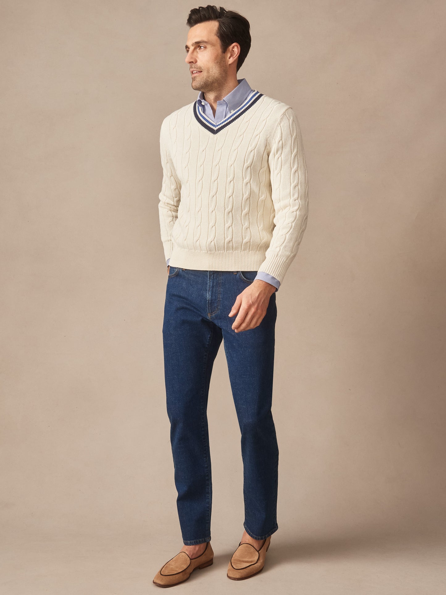 Man wearing an off-white Kent Sweater by J.McLaughlin, paired with a light blue shirt, dark jeans, and tan loafers, showcasing effortlessly stylish look with enduring designs.