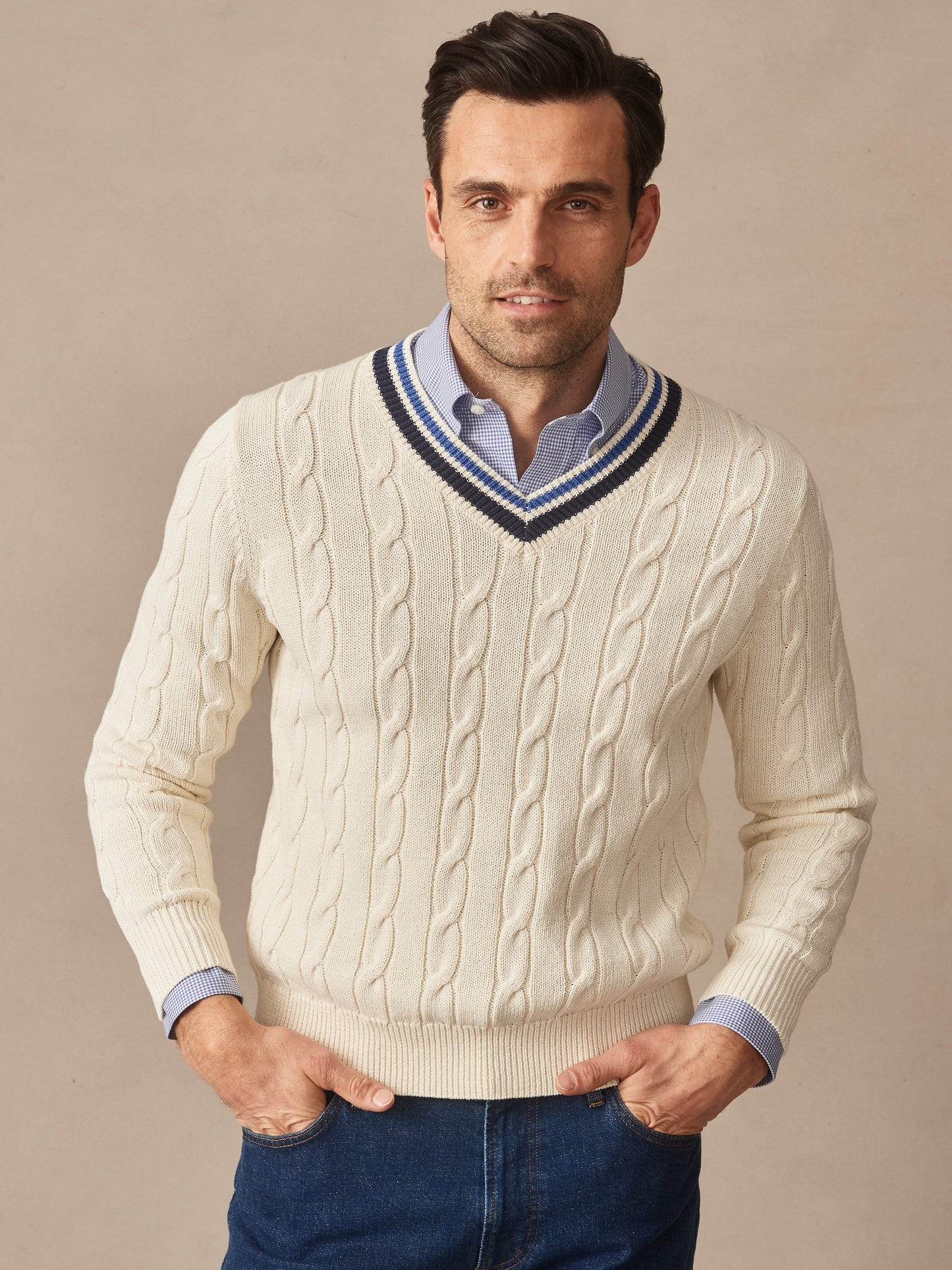 A man wearing a cream Kent Sweater by J.McLaughlin featuring a cable knit pattern and striped V-neck, paired with a blue shirt and jeans, showcasing effortlessly stylish and enduring designs.