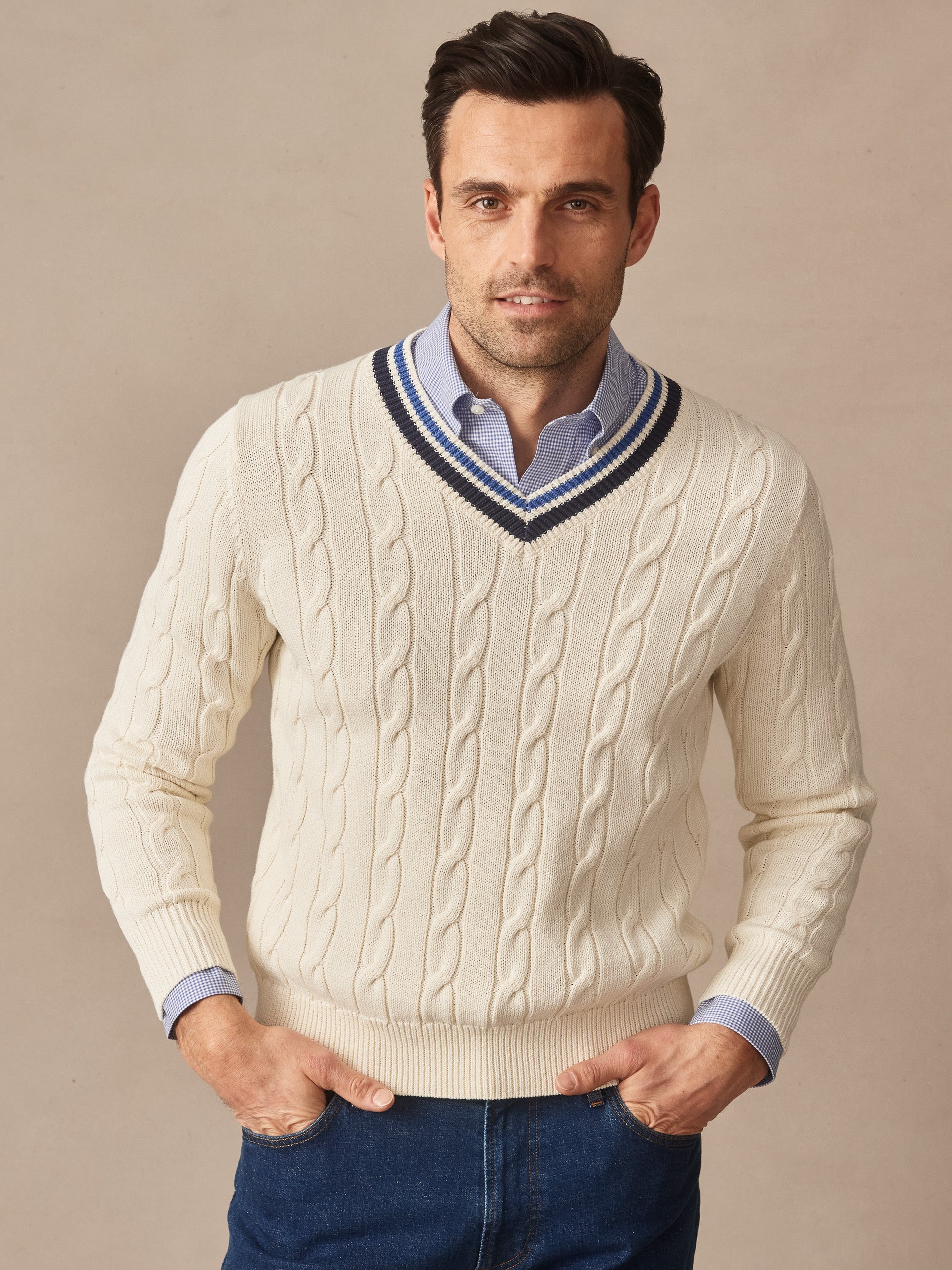 A man wearing a cream Kent Sweater by J.McLaughlin featuring a cable knit pattern and striped V-neck, paired with a blue shirt and jeans, showcasing effortlessly stylish and enduring designs.