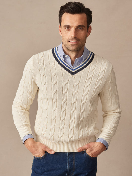 A man wearing a cream Kent Sweater by J.McLaughlin featuring a cable knit pattern and striped V-neck, paired with a blue shirt and jeans, showcasing effortlessly stylish and enduring designs.