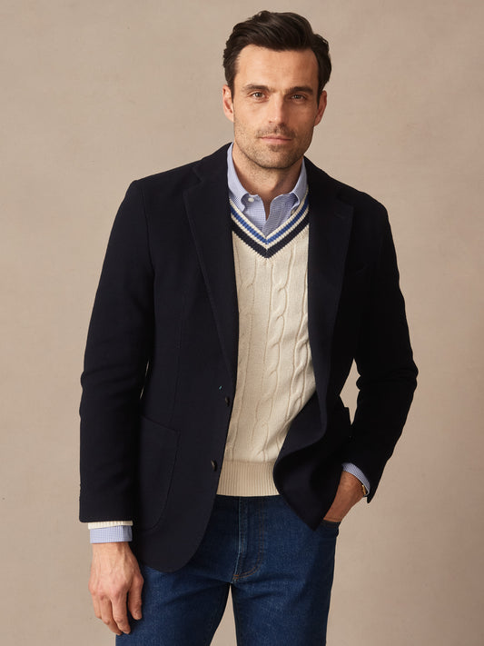 Man wearing a navy jacket over a cream cable-knit sweater with blue and white striped V-neck, paired with blue jeans, showcasing the Canary Knit Blazer by J.McLaughlin, effortlessly stylish and embodying enduring designs.