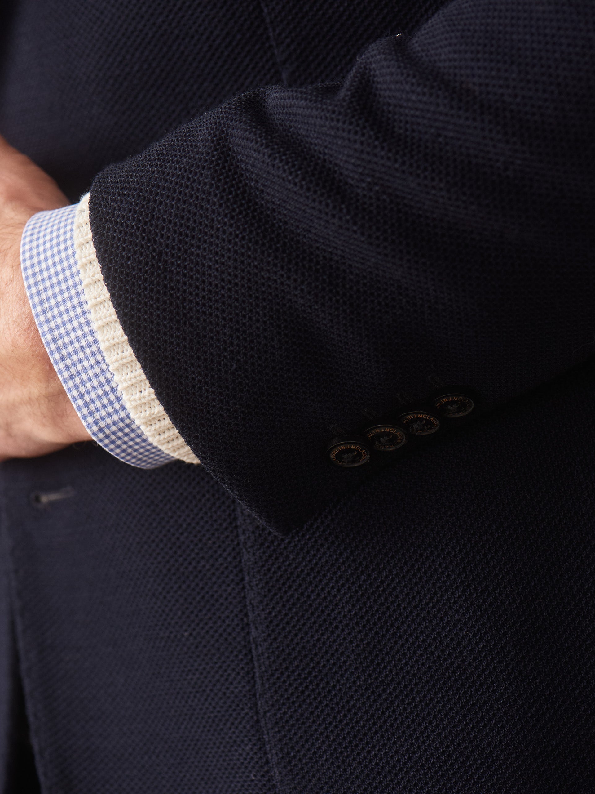 Close-up of a navy textured Canary Knit Blazer sleeve by J.McLaughlin, showcasing four buttons and layered over a light blue checkered shirt cuff with an off-white knit detail, reflecting effortlessly stylish, enduring designs in innovative fabrics from a destination for defining style. Life, Well-Lived.