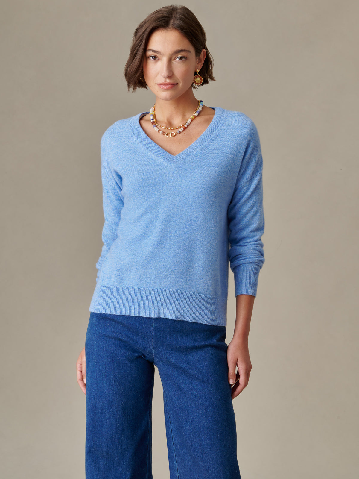 Woman wearing a light blue Kerry Cashmere Sweater by J.McLaughlin with a V-neck and ribbed trim, styled with layered necklaces and blue pants in a timeless, elegant look.