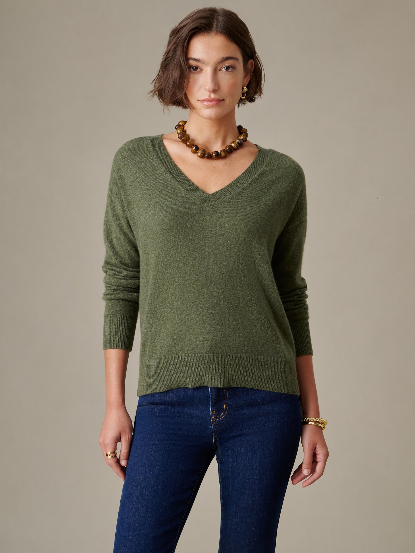 Woman wearing an olive green Kerry Cashmere Sweater by J.McLaughlin paired with dark blue jeans and accessorized with a beaded necklace and gold jewelry, capturing timeless style.