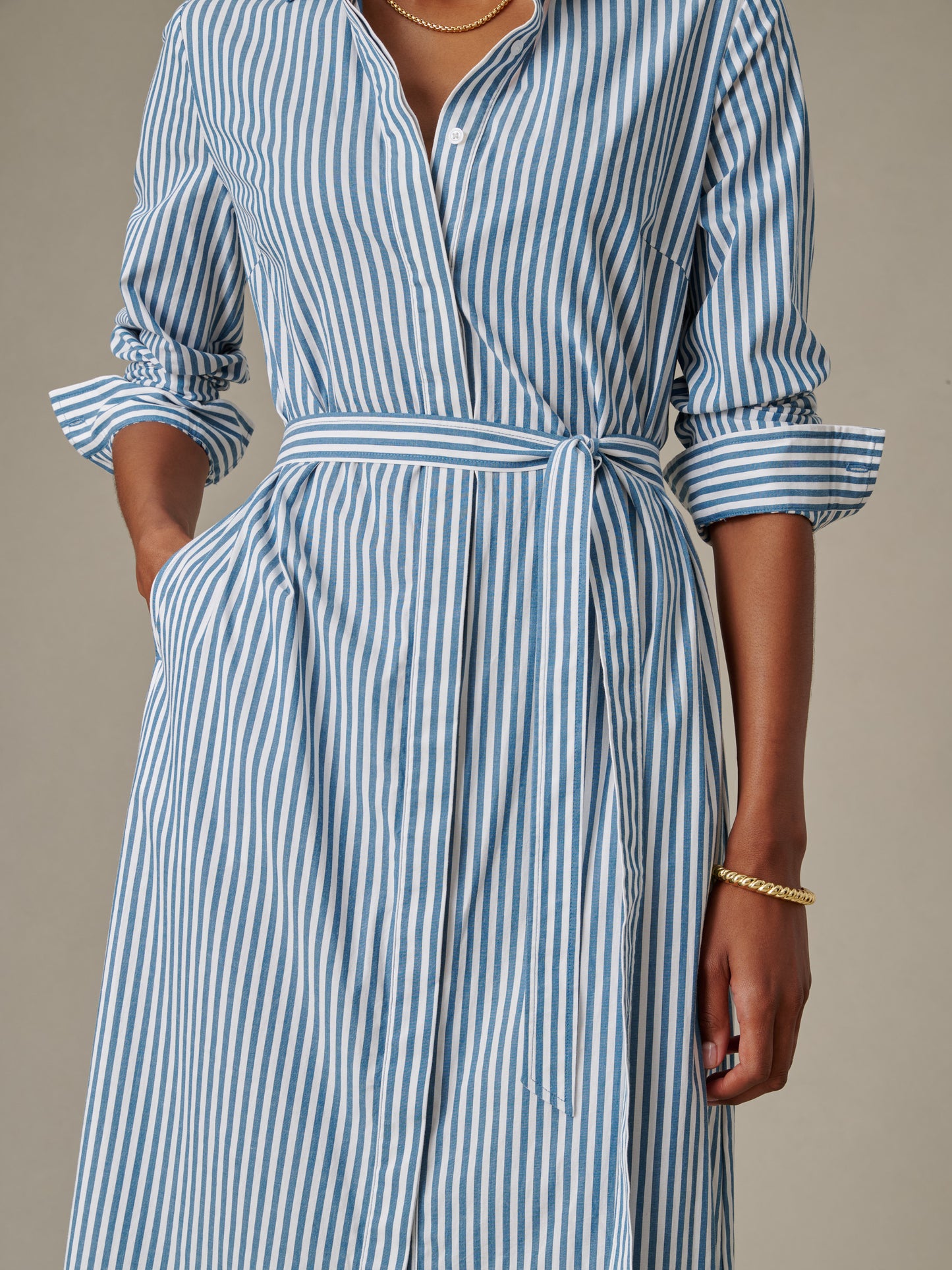 Model wearing a blue and white striped Leighton Dress in Stripe from J.McLaughlin with rolled sleeves, a tied waist belt, and gold jewelry accents.