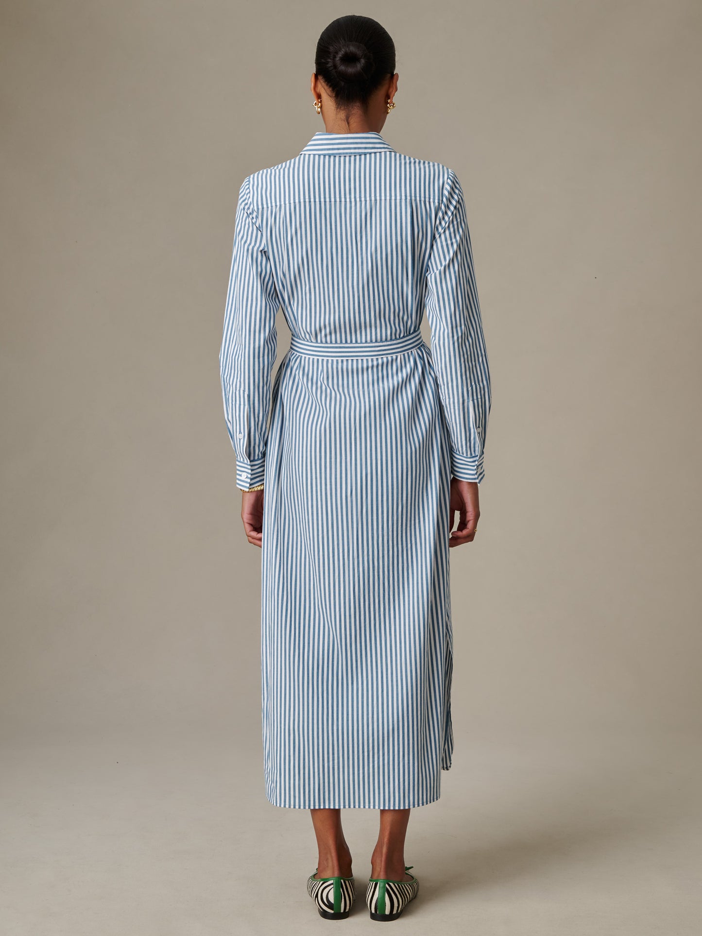 Woman wearing a blue and white striped Leighton Dress in Stripe by J.McLaughlin, featuring a belted waist and classic collar, paired with green and white shoes, standing against a neutral background.