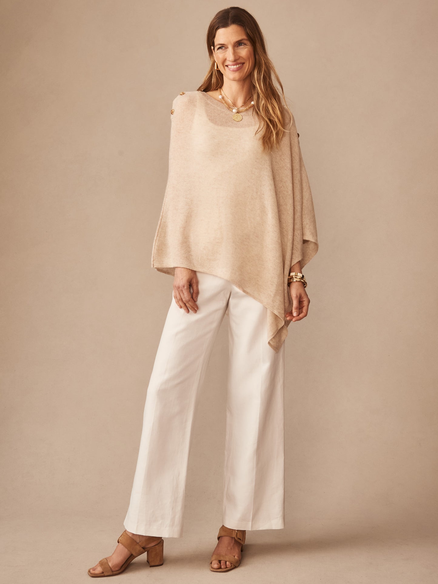 A woman models the Leila Cashmere Poncho by J.McLaughlin, featuring a tan, soft cashmere fabric with button details, paired with white wide-leg pants and tan sandals, embodying effortlessly stylish, enduring designs and innovative fabrics in a legacy of timeless fashion. A destination for defining style. Life, Well-Lived.