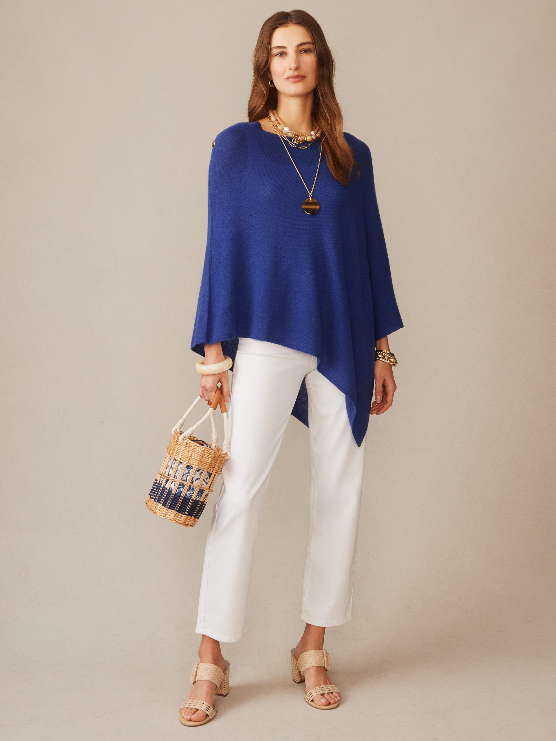 A woman modeling the Leila Cashmere Poncho by J.McLaughlin in a rich nautical navy tone, styled with white pants, tan sandals, layered jewelry, and a woven handbag, showcasing effortlessly stylish layering with enduring designs and innovative fabrics from a destination for defining style. Life, Well-Lived.