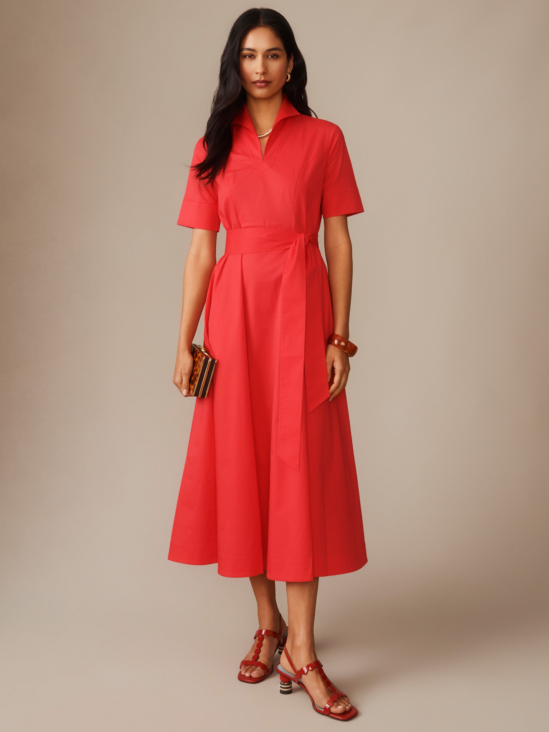 Model wearing a vibrant red Lily Dress by J.McLaughlin, featuring a collared neckline, short sleeves, and a tied waist belt, styled with matching sandals and a small clutch, perfectly blending effortlessly stylish, enduring designs and innovative fabrics.