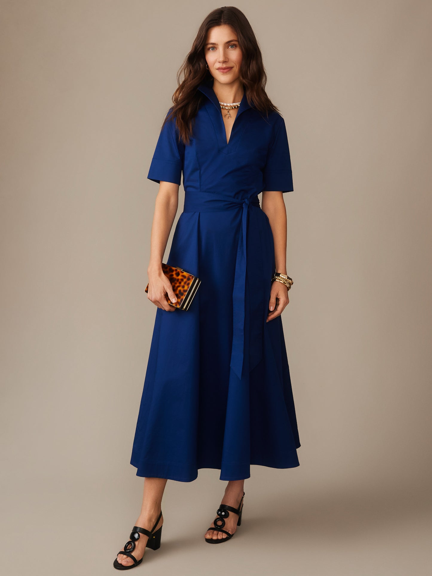 A woman wearing a nautical navy Lily Dress by J.McLaughlin, featuring a wrap design and short sleeves, styled with a tortoiseshell clutch and black heeled sandals, embodying effortlessly stylish, enduring designs within a legacy of timeless fashion. A destination for defining style. Life, Well-Lived.
