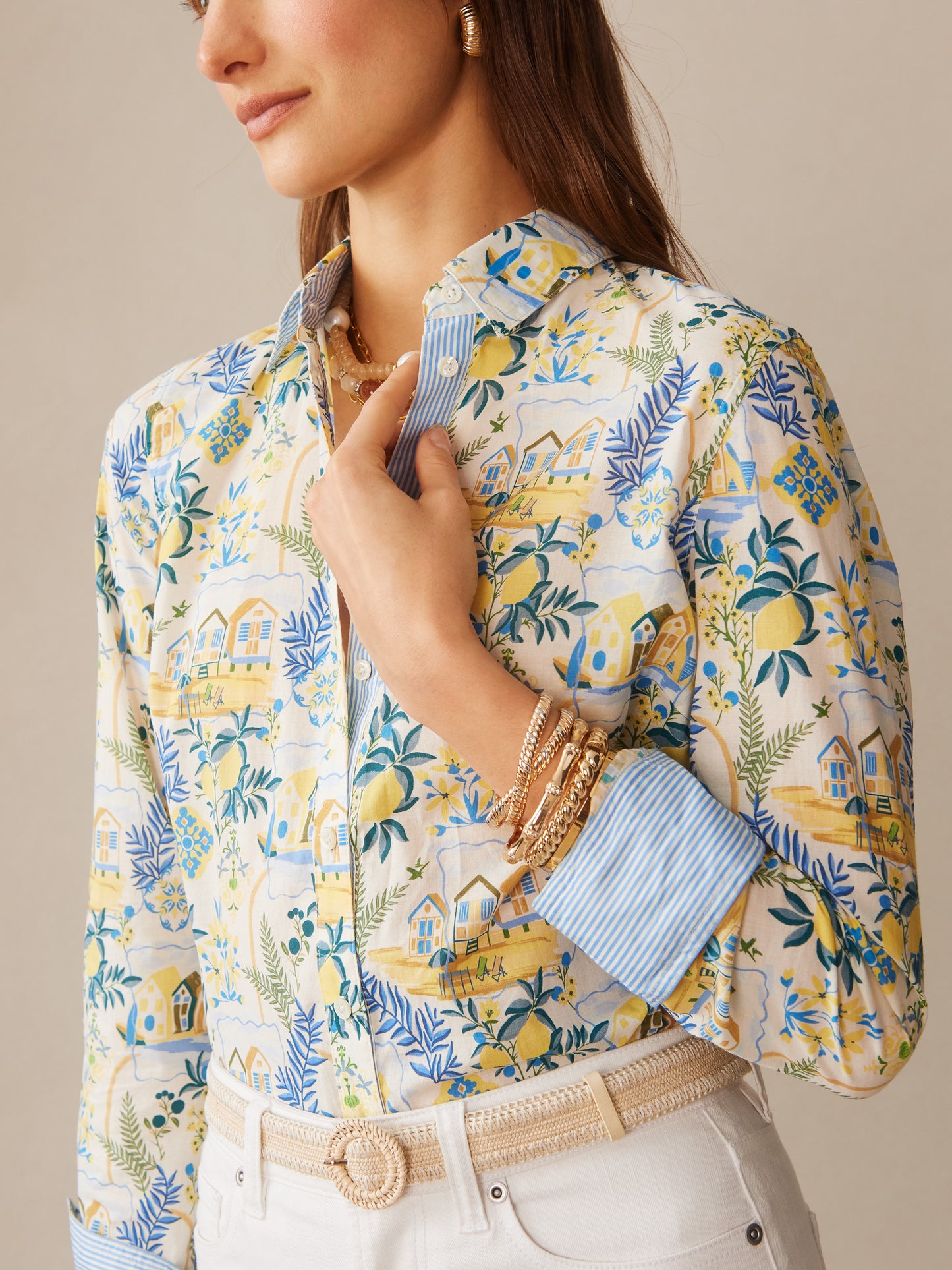 Woman wearing J.McLaughlin Lois Shirt in Mini Amalfi Coast Stripe, featuring an intricate coastal pattern with blues, yellows, and greens, paired with white pants and gold bracelets, showcasing effortlessly stylish, enduring designs and innovative fabrics. A destination for defining style. Life, Well-Lived.