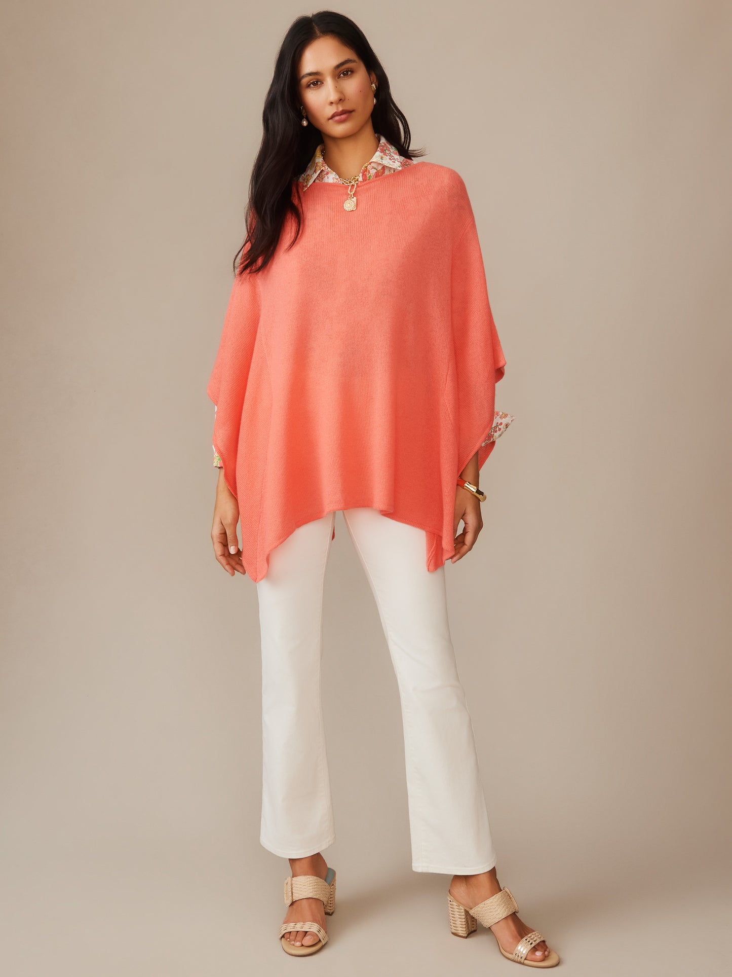 Woman wearing a coral Marin Cashmere Poncho by J.McLaughlin over a floral blouse paired with white pants and beige heeled sandals, showcasing effortlessly stylish, enduring designs and innovative fabrics.