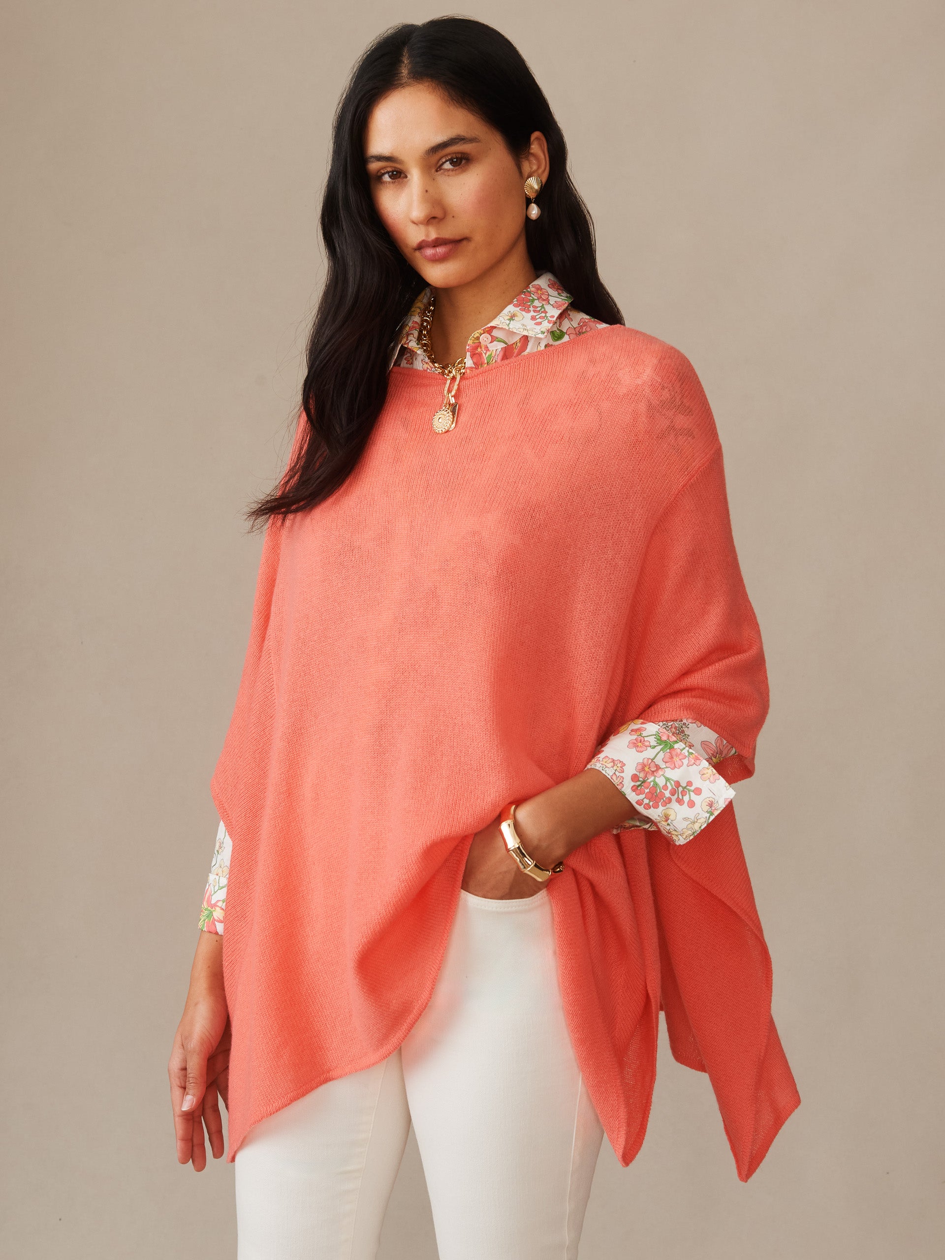 A woman wearing a coral Marin Cashmere Poncho layered over a floral blouse and white pants, showcasing J.McLaughlin’s effortlessly stylish, enduring designs in timeless fashion.