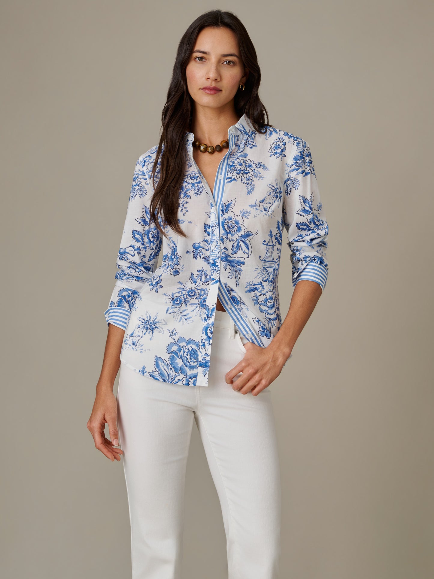 Woman wearing the Lois Shirt in Antique Toile Stripe by J.McLaughlin, featuring a classic blue and white floral pattern with striped inner cuffs, styled with white pants and a bold necklace, showcasing effortless style and timeless elegance.