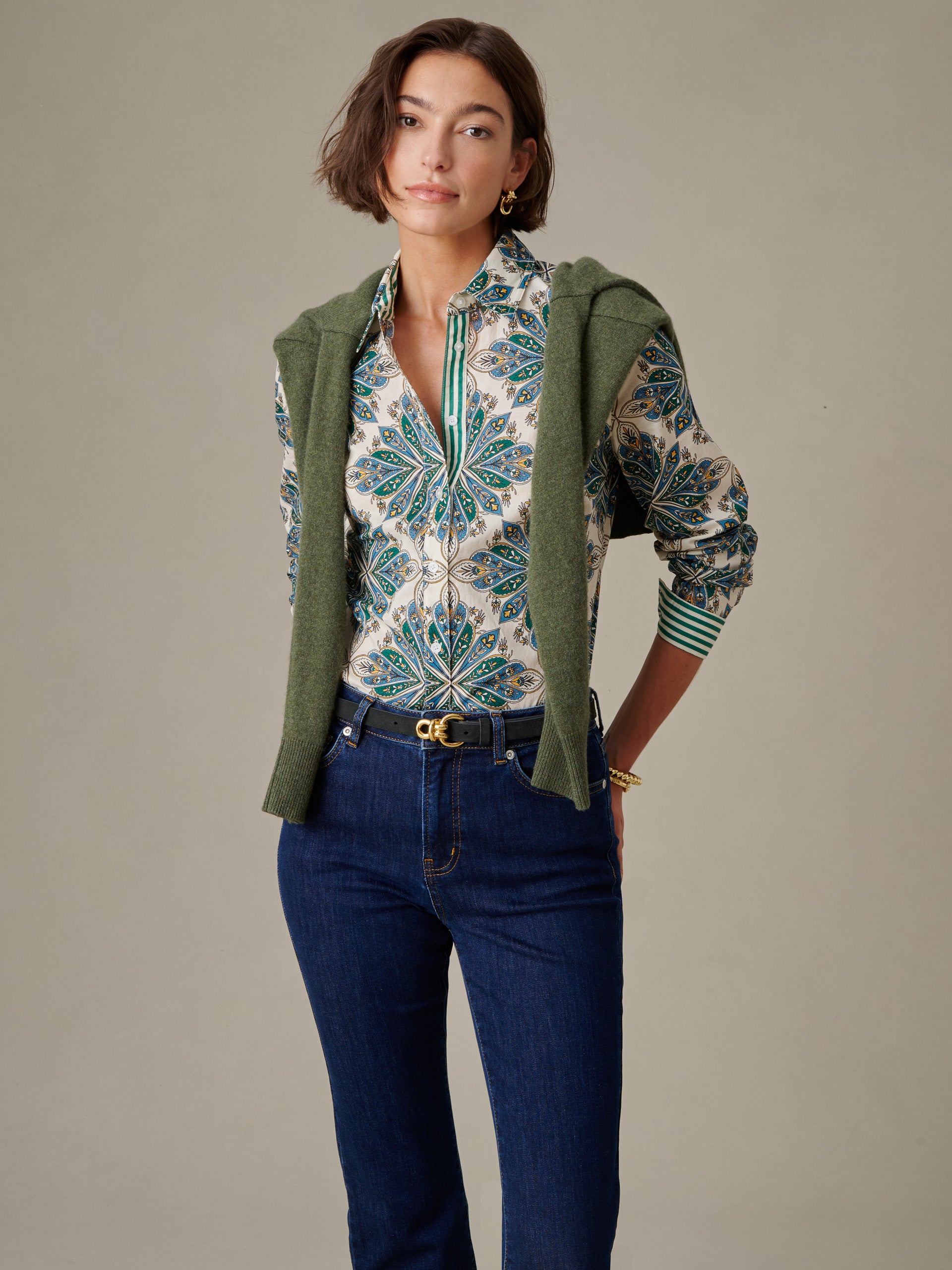 Woman wearing a patterned Lois Shirt in Neo Radiant Stripe by J.McLaughlin paired with dark jeans and an olive green sweater draped over her shoulders, showcasing a timeless, effortlessly stylish look.