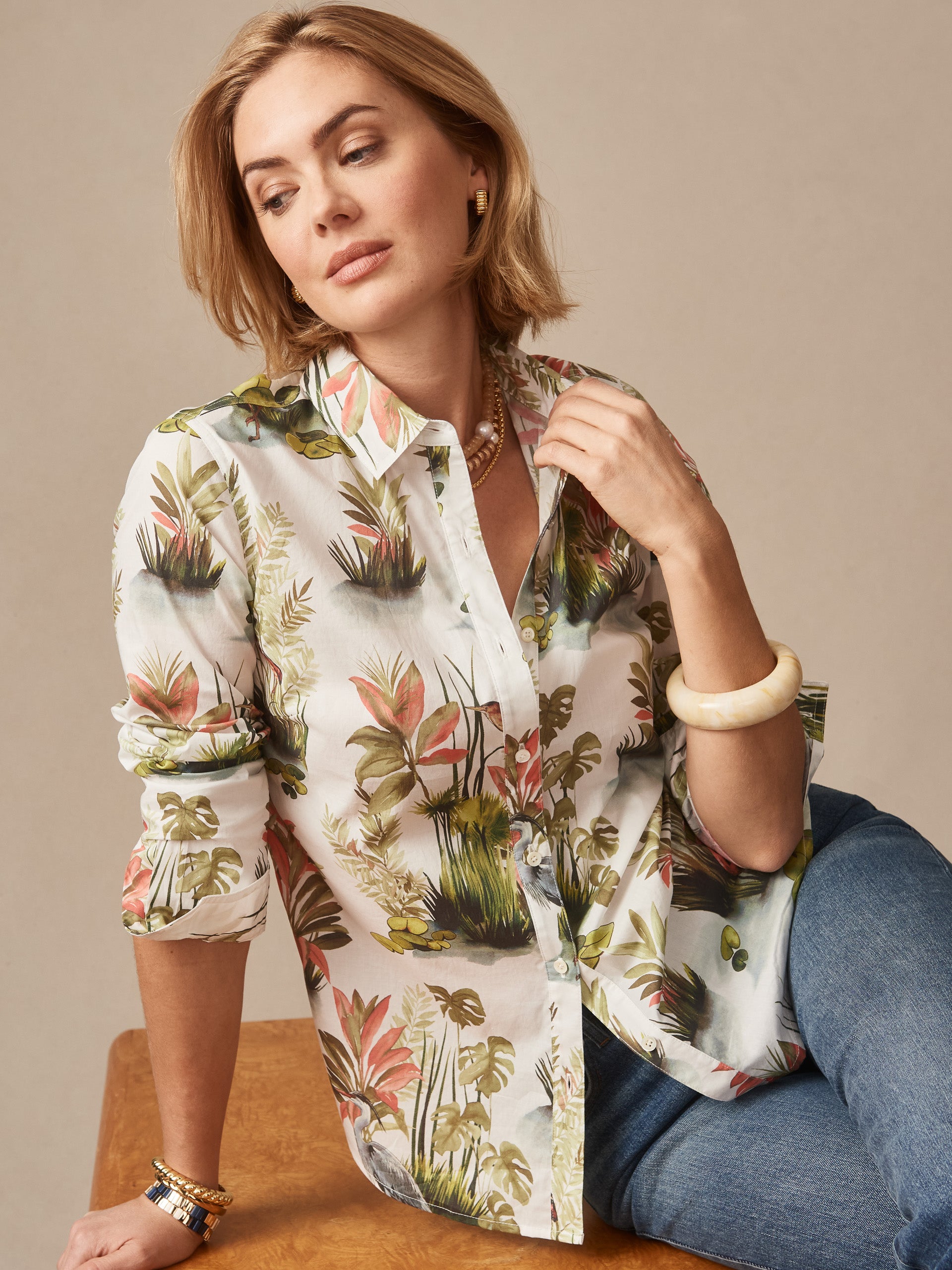 Woman wearing the Lois Shirt in Tidal Palms by J.McLaughlin, featuring effortlessly stylish tropical print blouse in soft coral, green, and white tones with rolled sleeves, paired with jeans, embodying enduring designs and innovative fabrics for a legacy of timeless fashion. A destination for defining style. Life, Well-Lived.