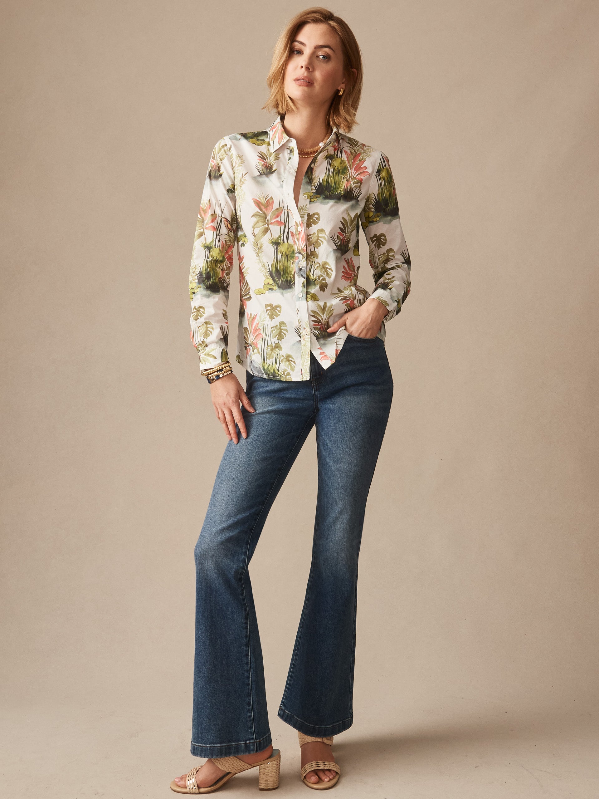 Woman wearing J.McLaughlin Lois Shirt in Tidal Palms with floral print paired with flared jeans and sandals, showcasing effortlessly stylish, enduring designs and a legacy of timeless fashion.