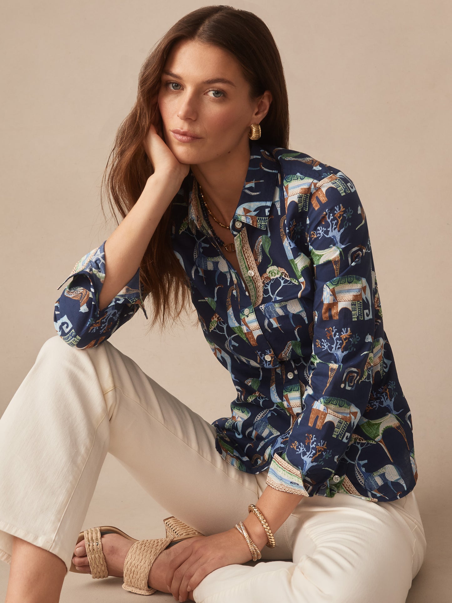 A woman wearing a J.McLaughlin Lois Shirt in The Wild Stripe with a colorful animal and nature pattern, paired with white pants and woven sandals, showcasing effortlessly stylish, enduring designs and innovative fabrics from a destination for defining style. Life, Well-Lived.