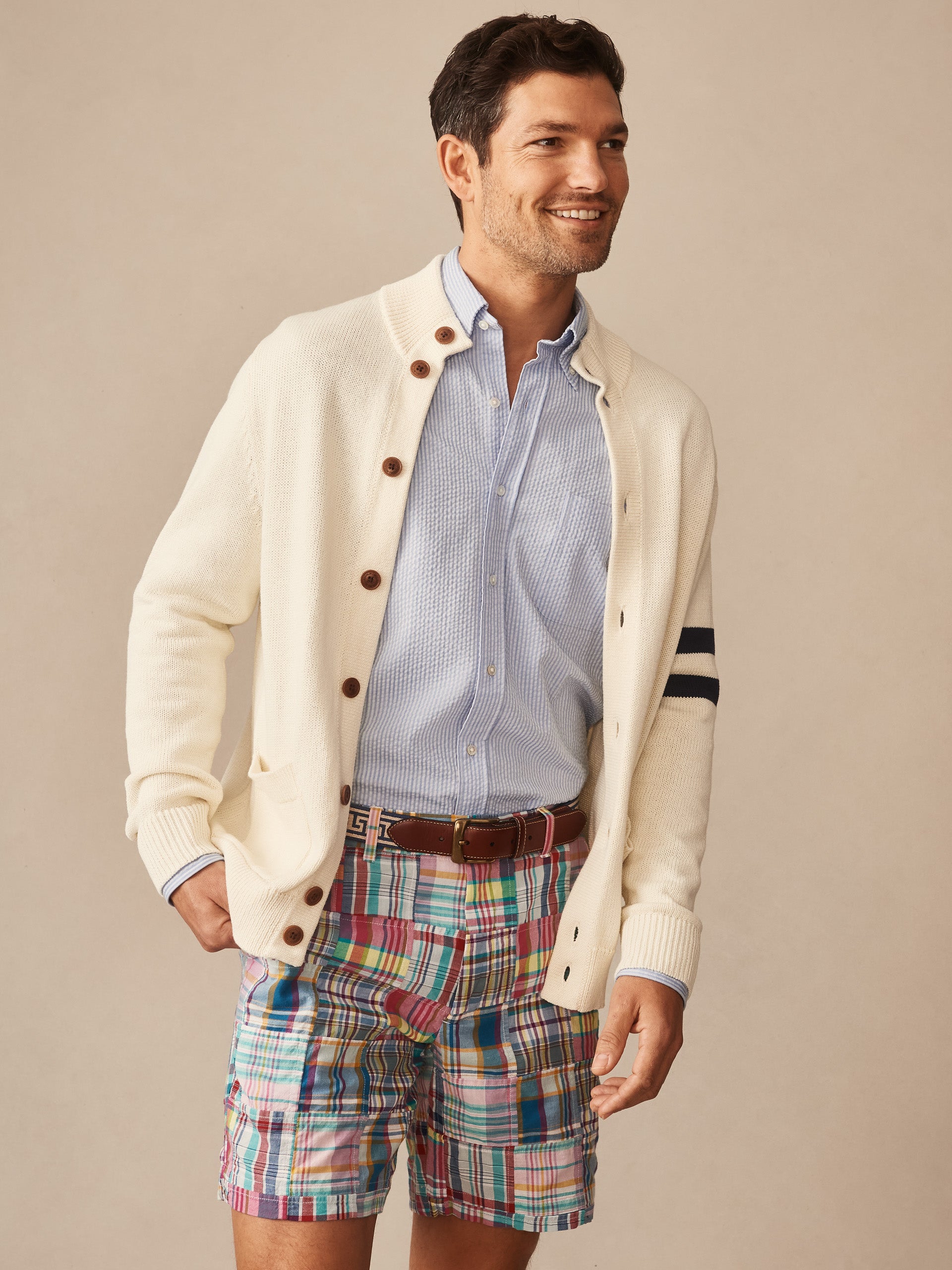 Man wearing an off-white Lowell Sweater from J.McLaughlin over a light blue button-down shirt paired with colorful plaid shorts, smiling and standing against a neutral background.