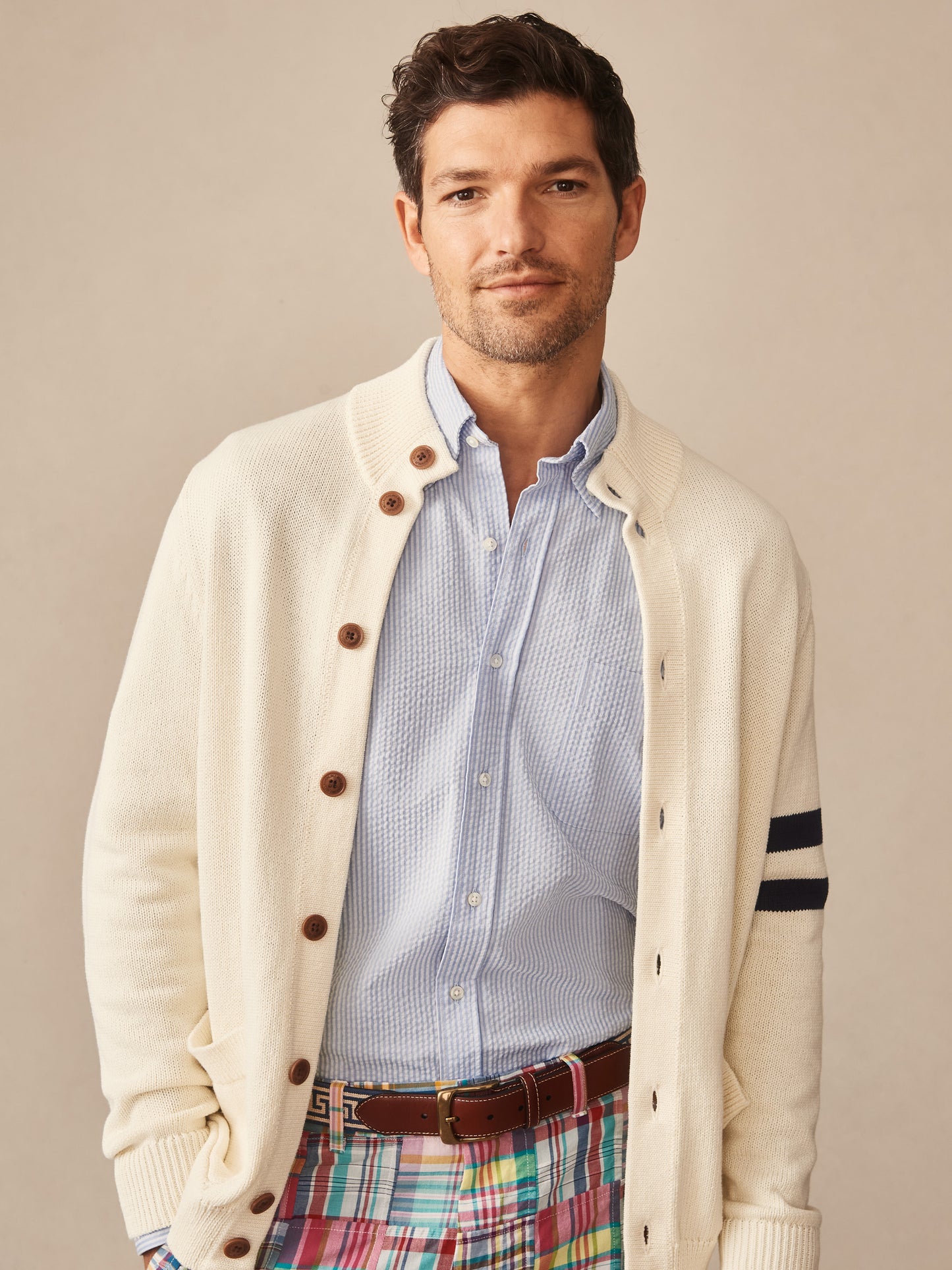 Man wearing an off-white Lowell Sweater by J.McLaughlin with brown buttons and navy stripes on the sleeve, layered over a light blue textured shirt and colorful plaid pants.