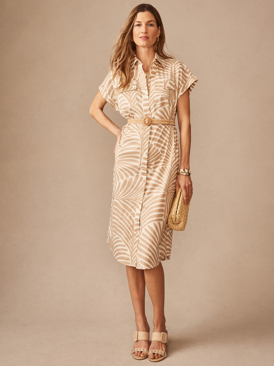 A woman wearing the Luciana Dress in Mod Frond by J.McLaughlin, featuring a tan and off-white palm leaf pattern, accessorized with a woven belt and clutch, epitomizing effortlessly stylish, enduring designs in innovative fabrics for a legacy of timeless fashion. A destination for defining style. Life, Well-Lived.