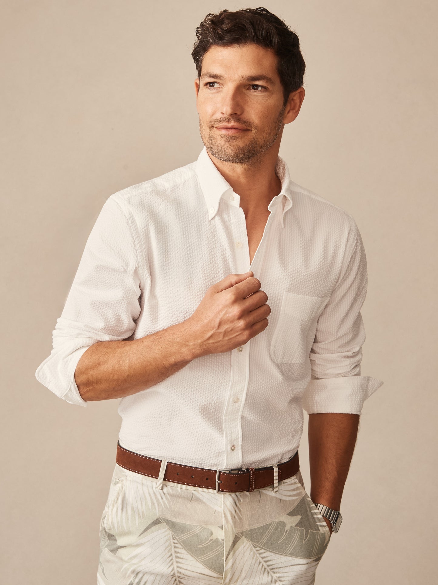 A man wearing a white Belvedere Cotton Seersucker Shirt by J.McLaughlin, styled with a brown belt and tropical pattern pants, showcasing effortlessly stylish, enduring designs with innovative fabrics in a legacy of timeless fashion. A destination for defining style. Life, Well-Lived.